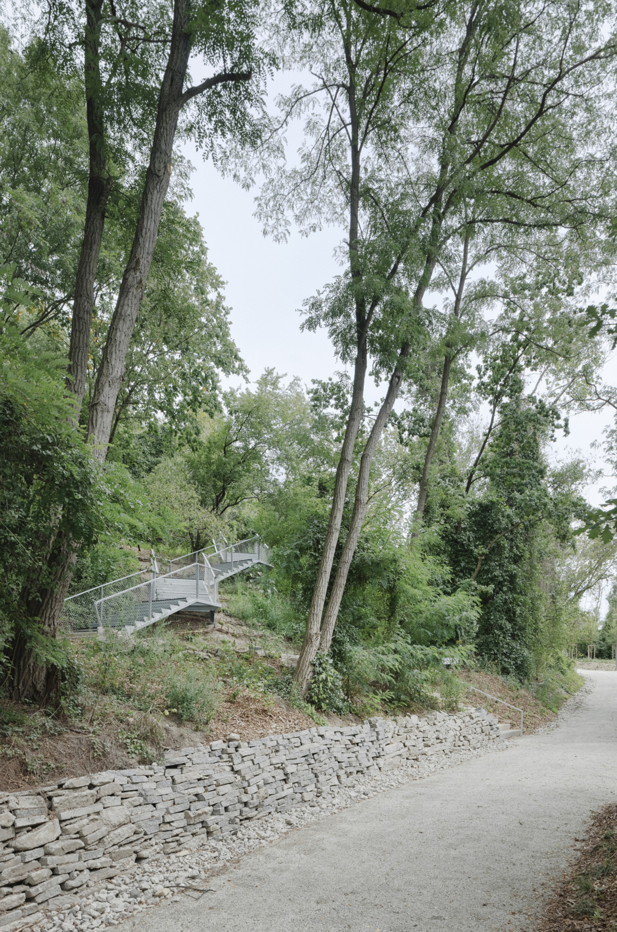 Metal footbridge ascending hillside amid trees beside a stone gabion wall and paved path