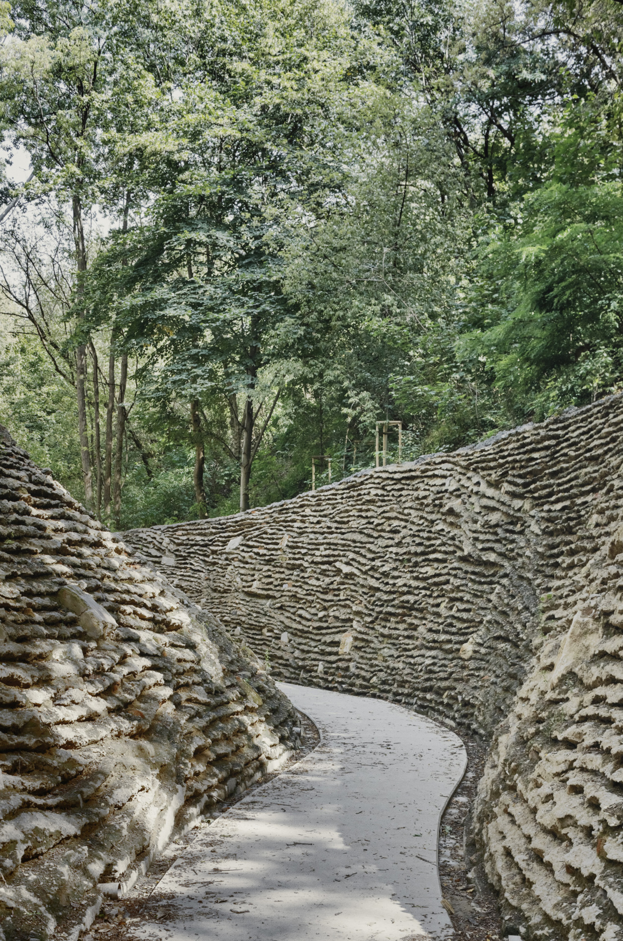 Curving pathway enclosed by textured stone walls with rippled horizontal patterns beneath a canopy of trees