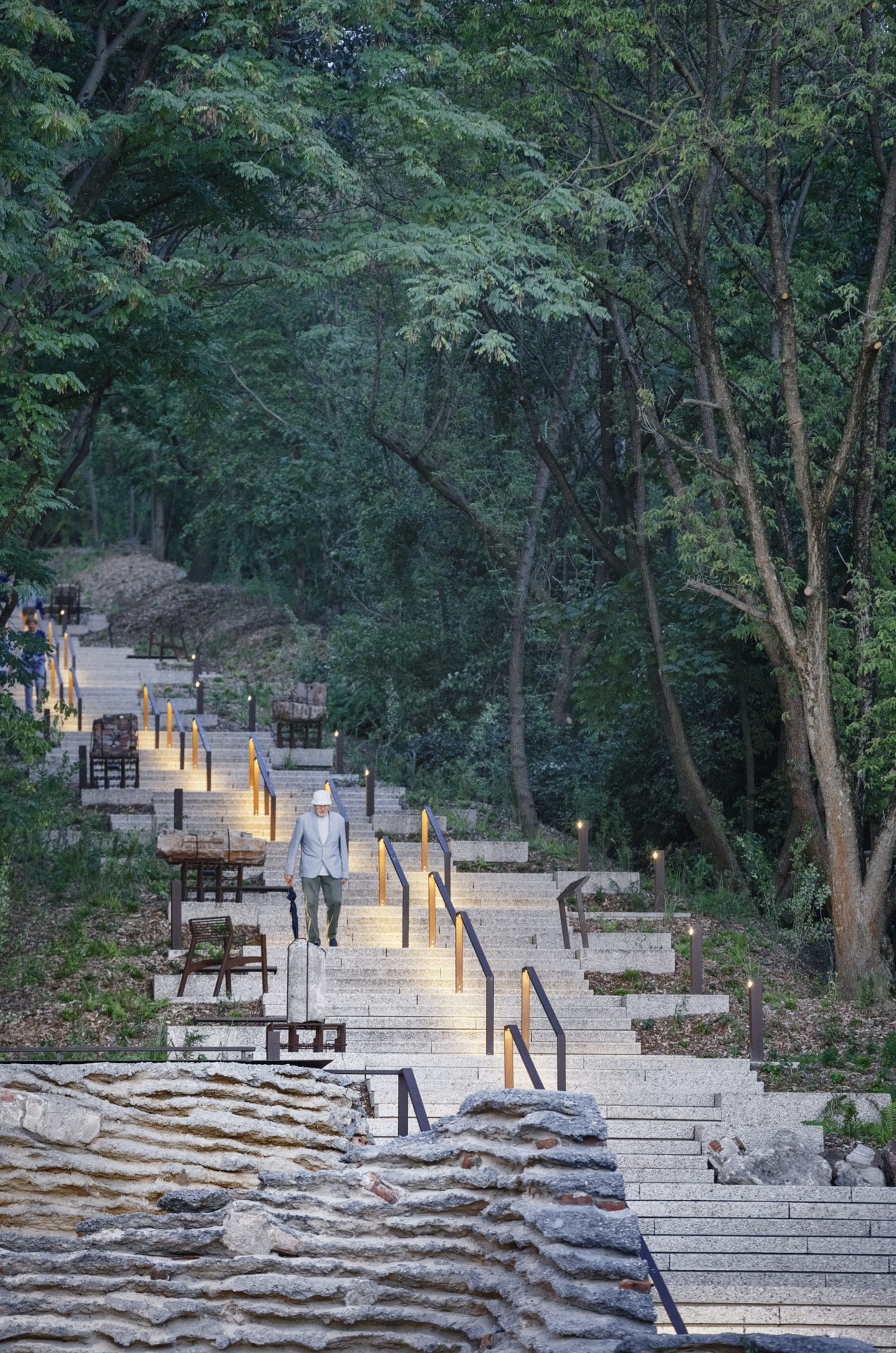 Timber boardwalk with handrails and lighting descending through forest at twilight with visitors walking