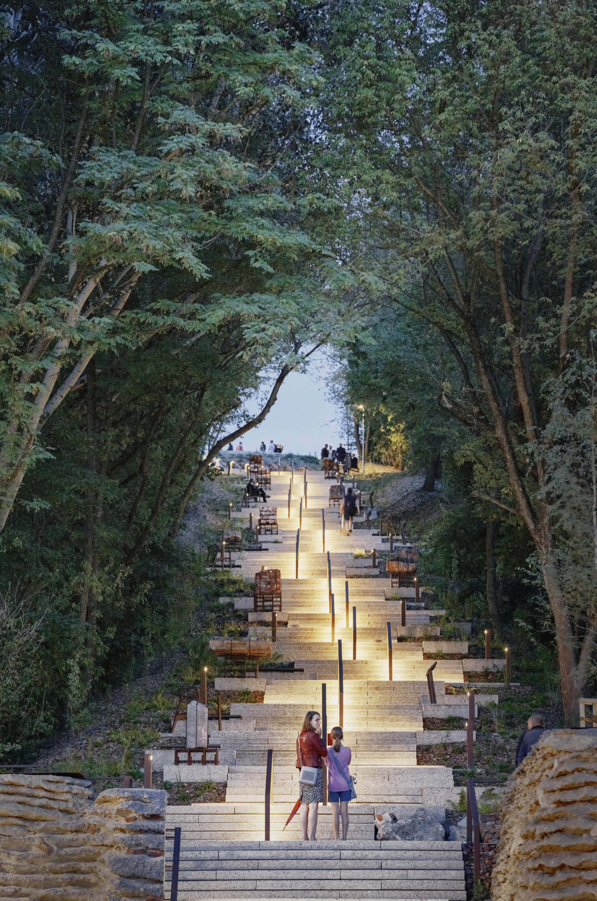 Illuminated stone stairway ascending through a canopy of trees at dusk with visitors scattered along the path