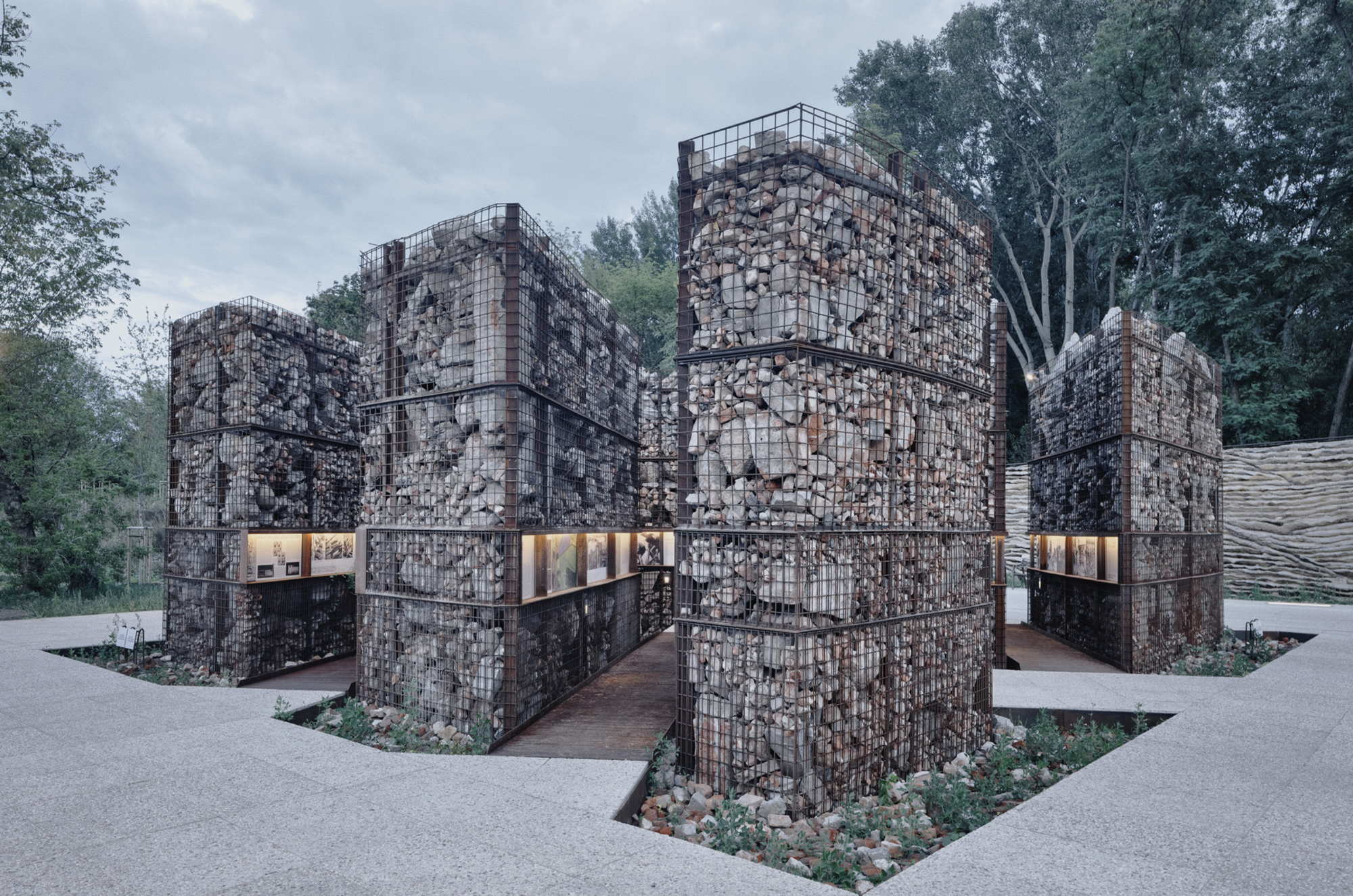 Cluster of cubic gabion volumes filled with rubble stone and integrated horizontal window openings at dusk