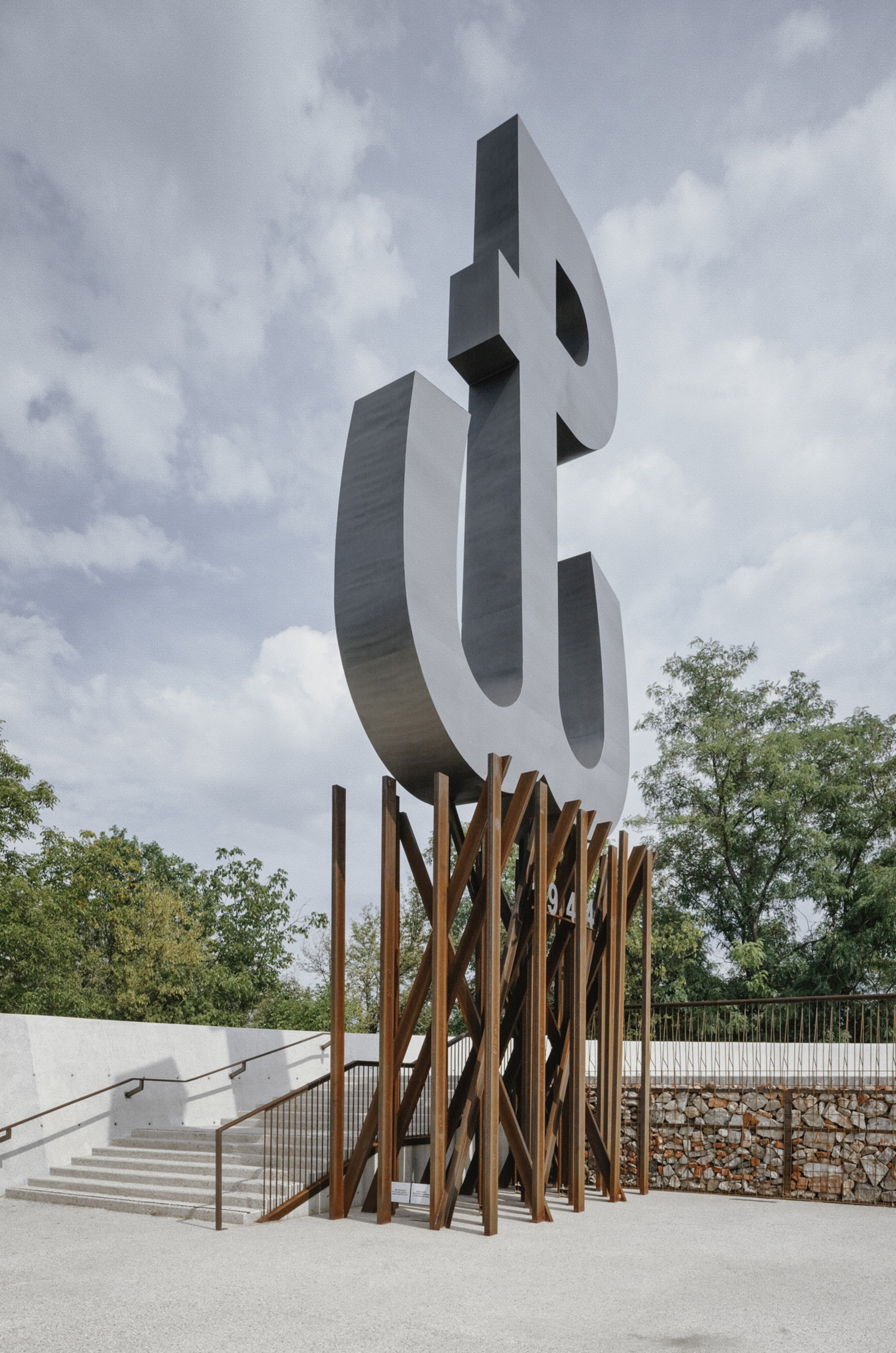 Sculptural metal letterform monument on vertical timber base beside concrete ramp and gabion retaining wall under cloudy sky