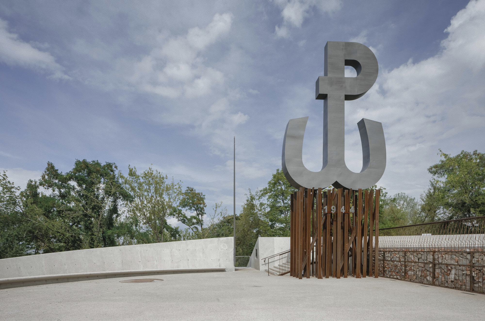 Metal letterform sculpture mounted on weathered timber slat screen beside concrete plaza and gabion wall with trees beyond