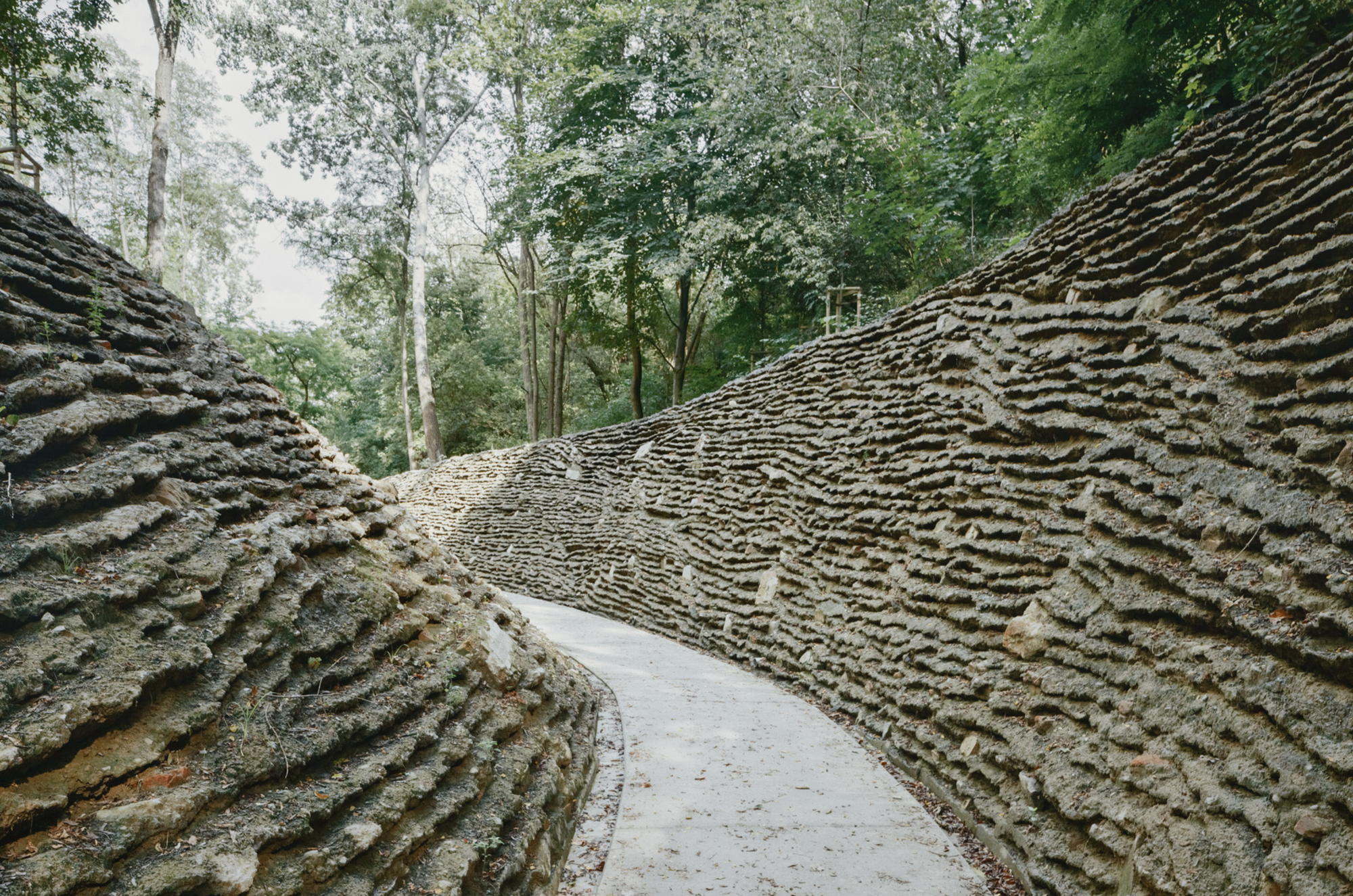 Curved concrete walkway enclosed by textured stone retaining walls with horizontal stratified layering under forest canopy