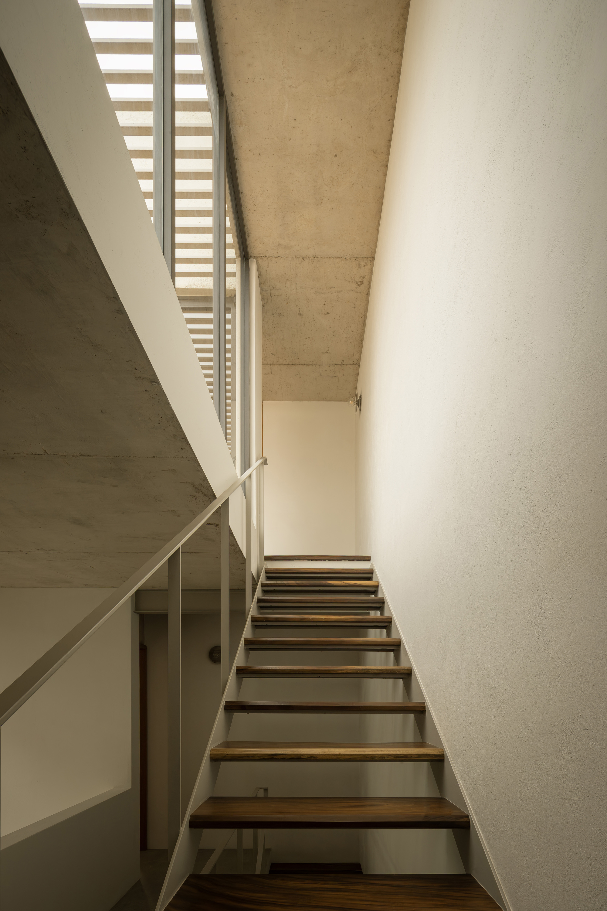 Stairwell with open timber treads and steel stringers ascending beneath a concrete soffit and skylight