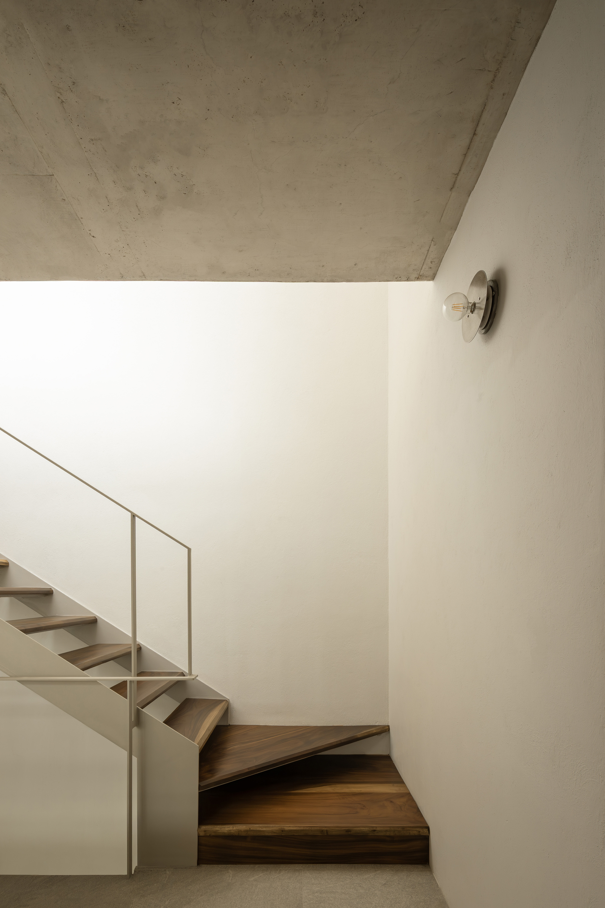 Wood staircase turning upward along white wall with circular sconce and concrete soffit above
