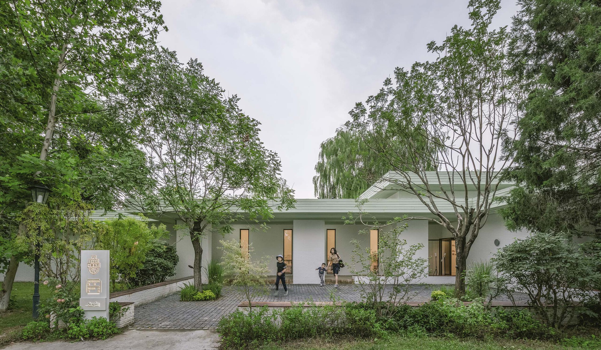 Front facade with vertical window openings and brick paving framed by mature trees under an overcast sky