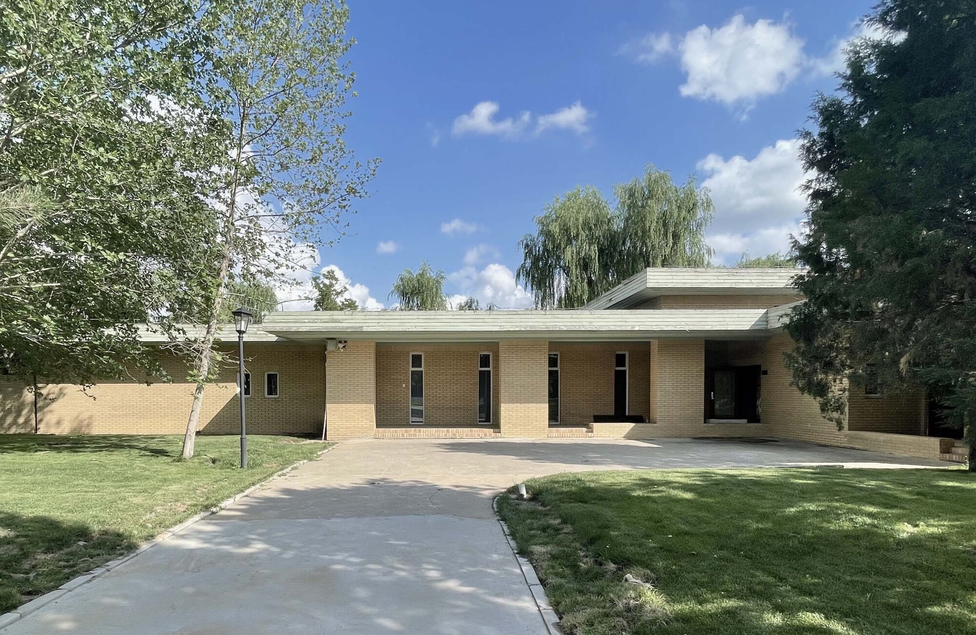 Front facade of buff brick with flat roof and tall willow trees framing the lawn