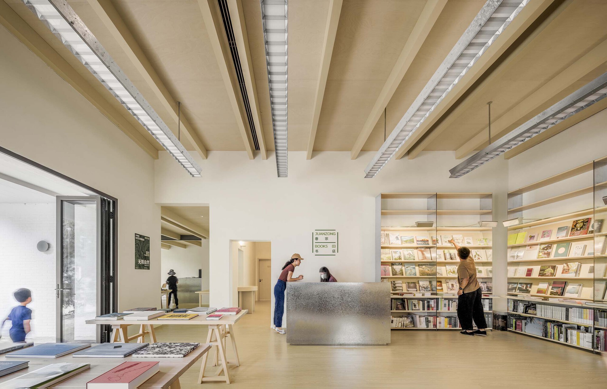 Reception area with metal desk and plywood bookshelves beneath exposed ceiling beams and linear lighting