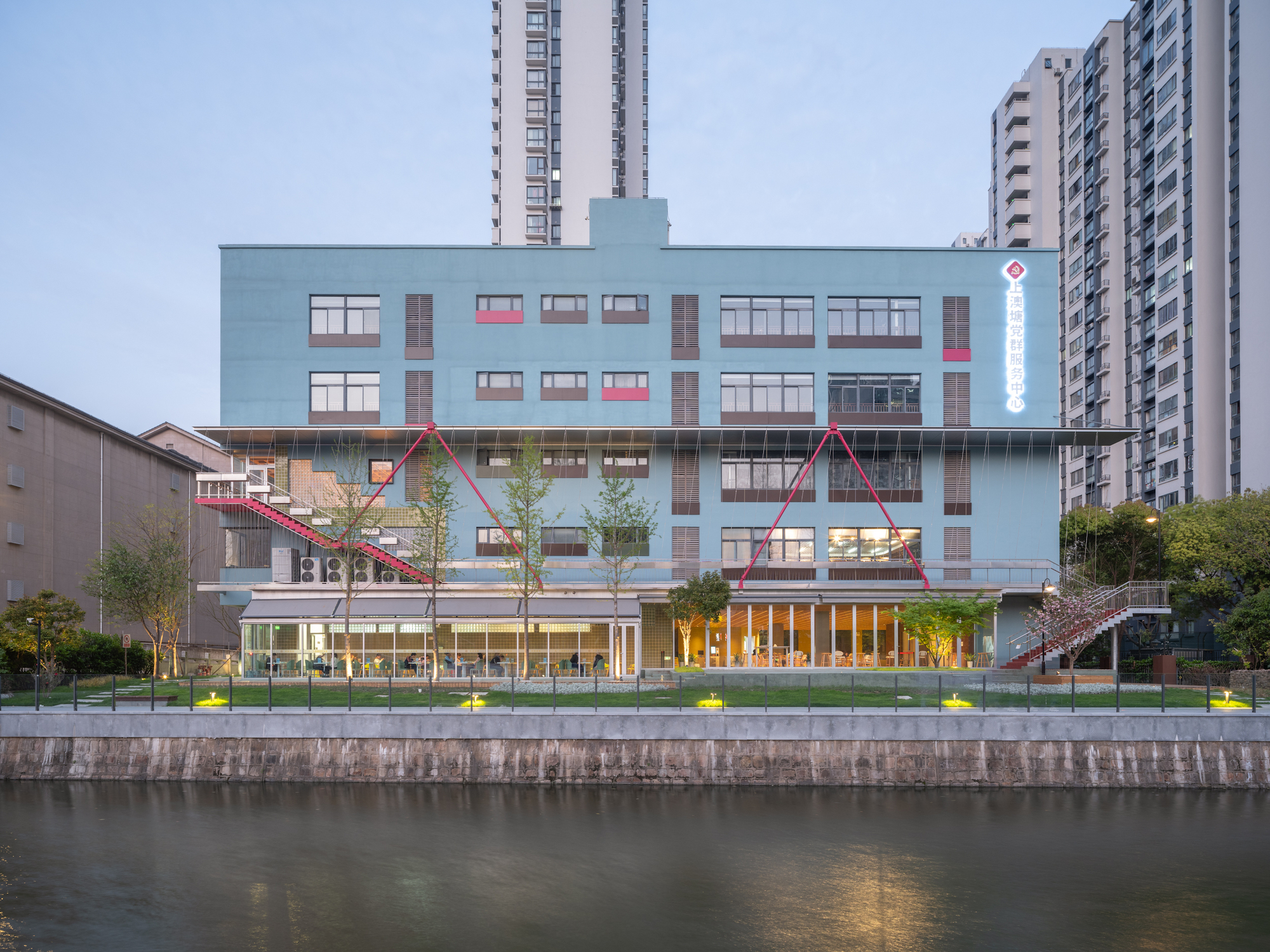 Blue facade with punched windows and triangular pink canopies viewed across the canal at dusk