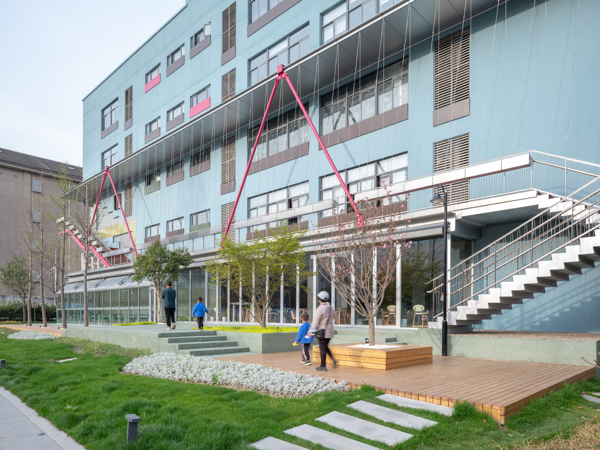 Ground-level courtyard with timber deck, planted lawn, and pink steel canopy structures with visitors passing