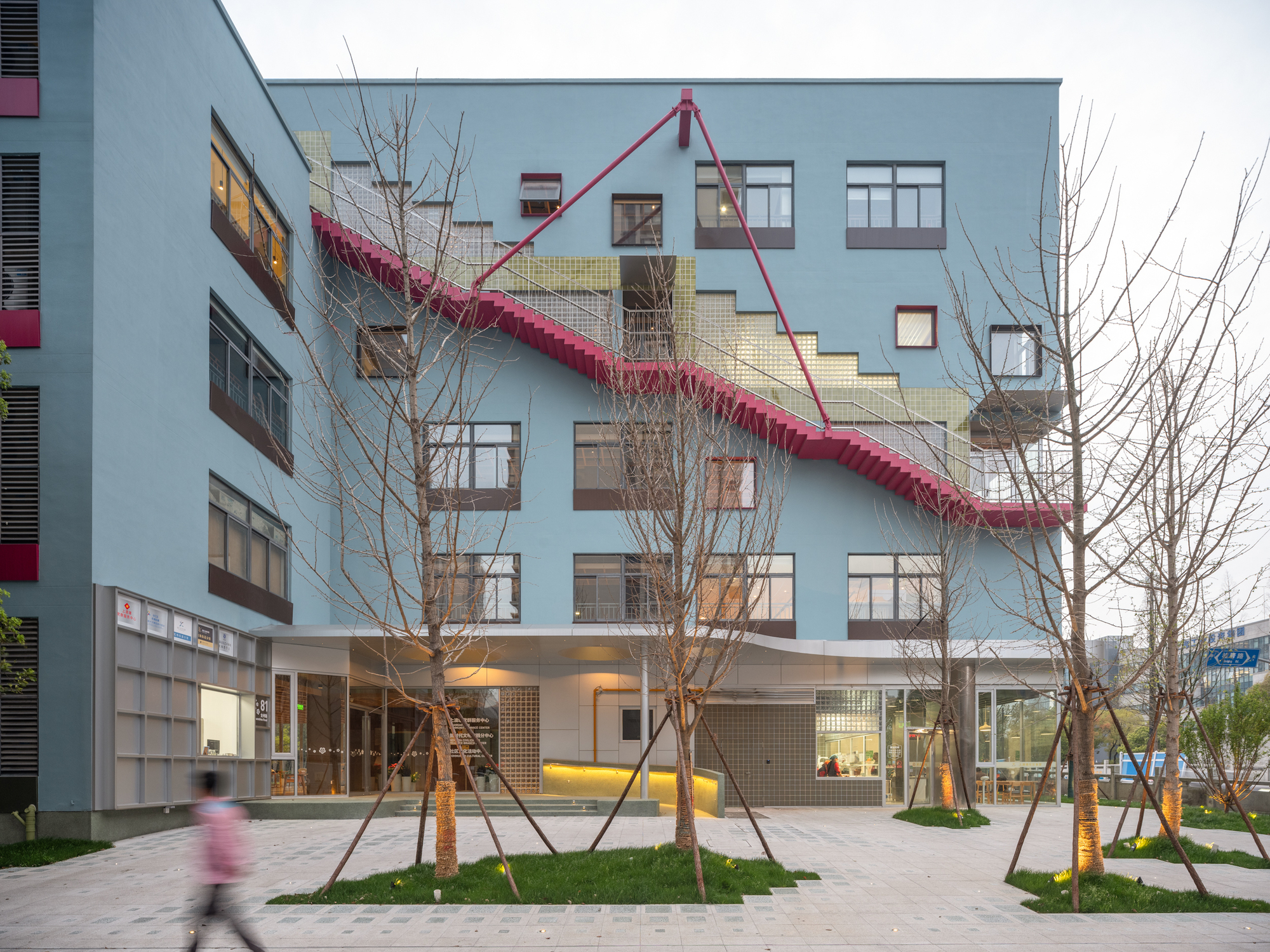 Street view of the pale blue facade with diagonal pink staircase and newly planted trees at dusk