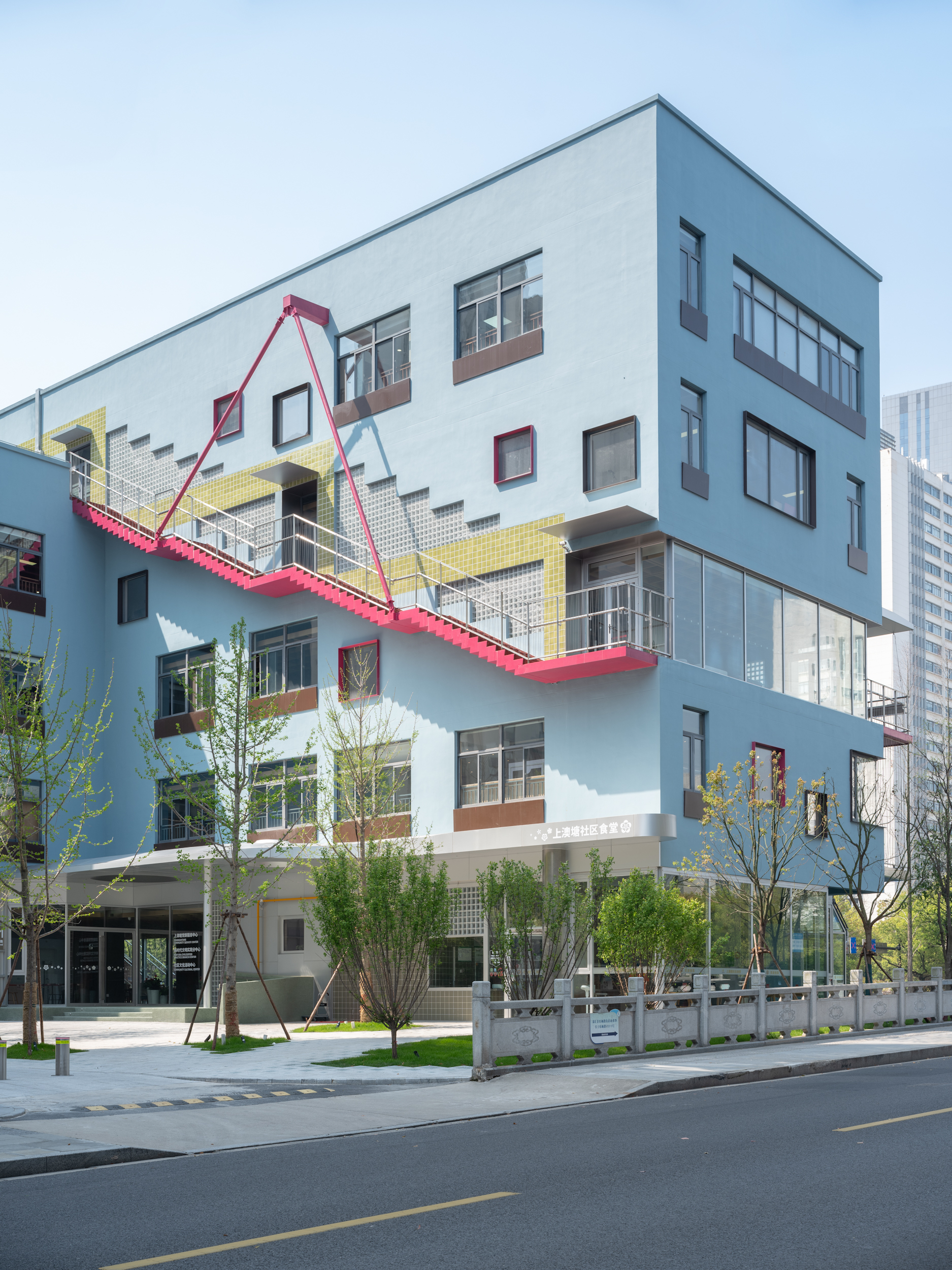 Corner facade showing the pale blue cladding, yellow tile band, and pink steel staircase with young street trees