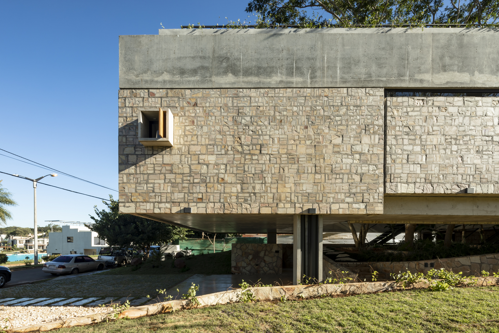 Street view of the stone clad facade with concrete beam and pilotis beneath clear blue sky