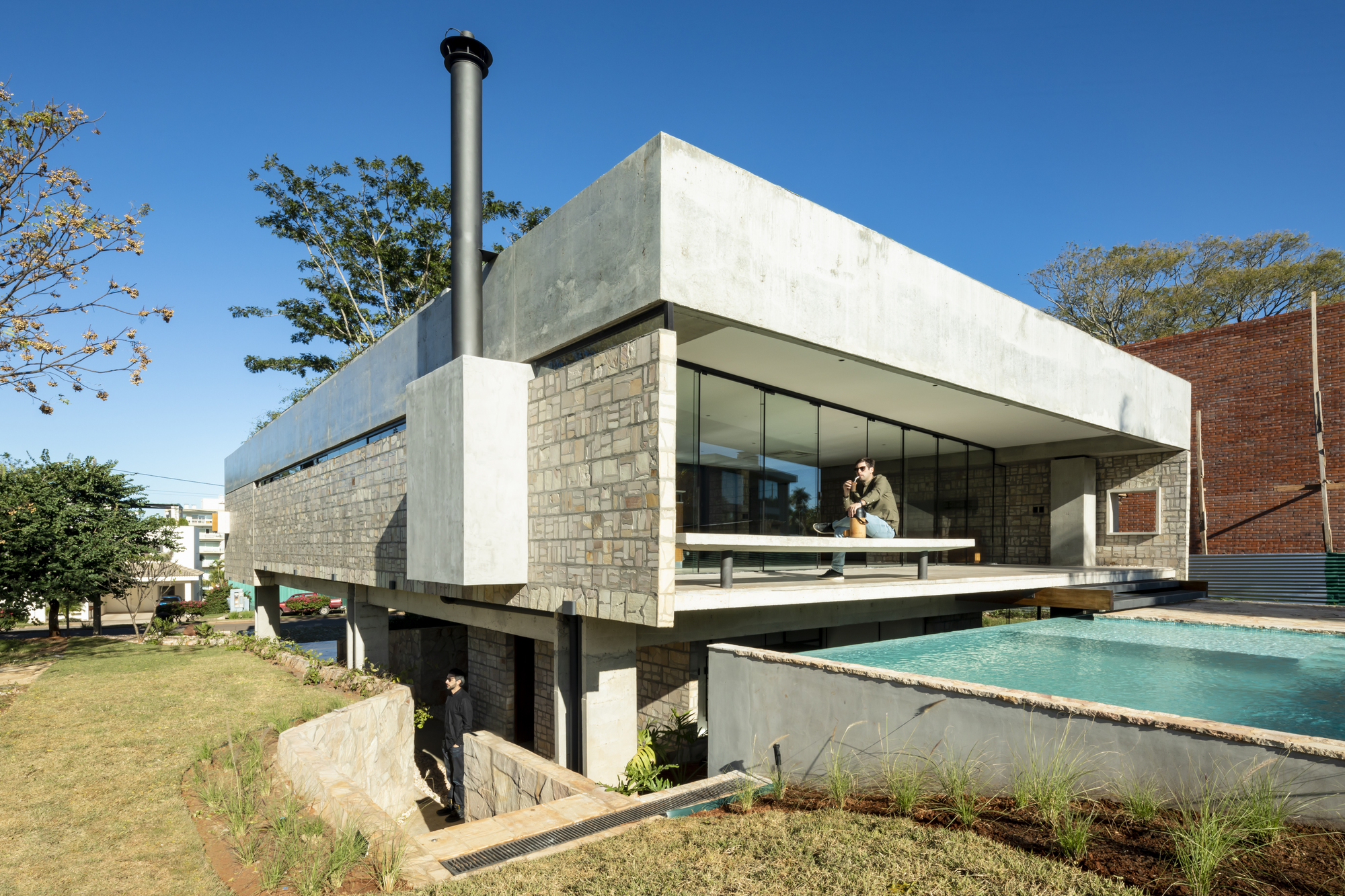 Corner view showing cantilevered concrete volume above concrete block base with swimming pool and two figures