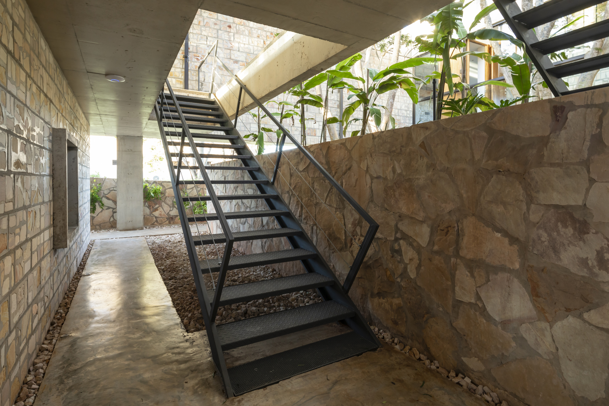 Open steel staircase ascending between stone walls and concrete ceiling with planted courtyard below