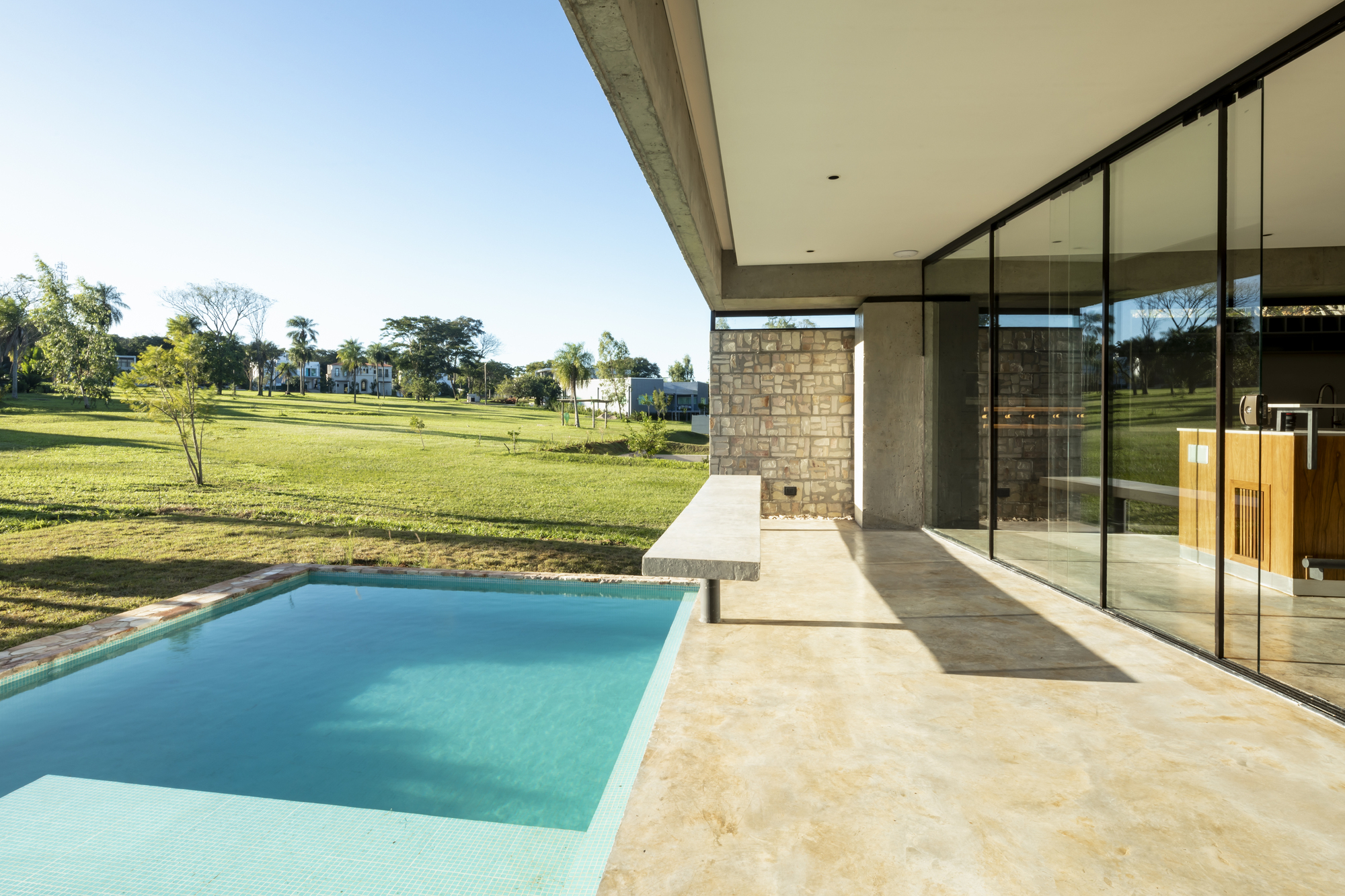 Covered terrace with limestone pavers overlooking a rectangular pool and lawn under clear blue sky