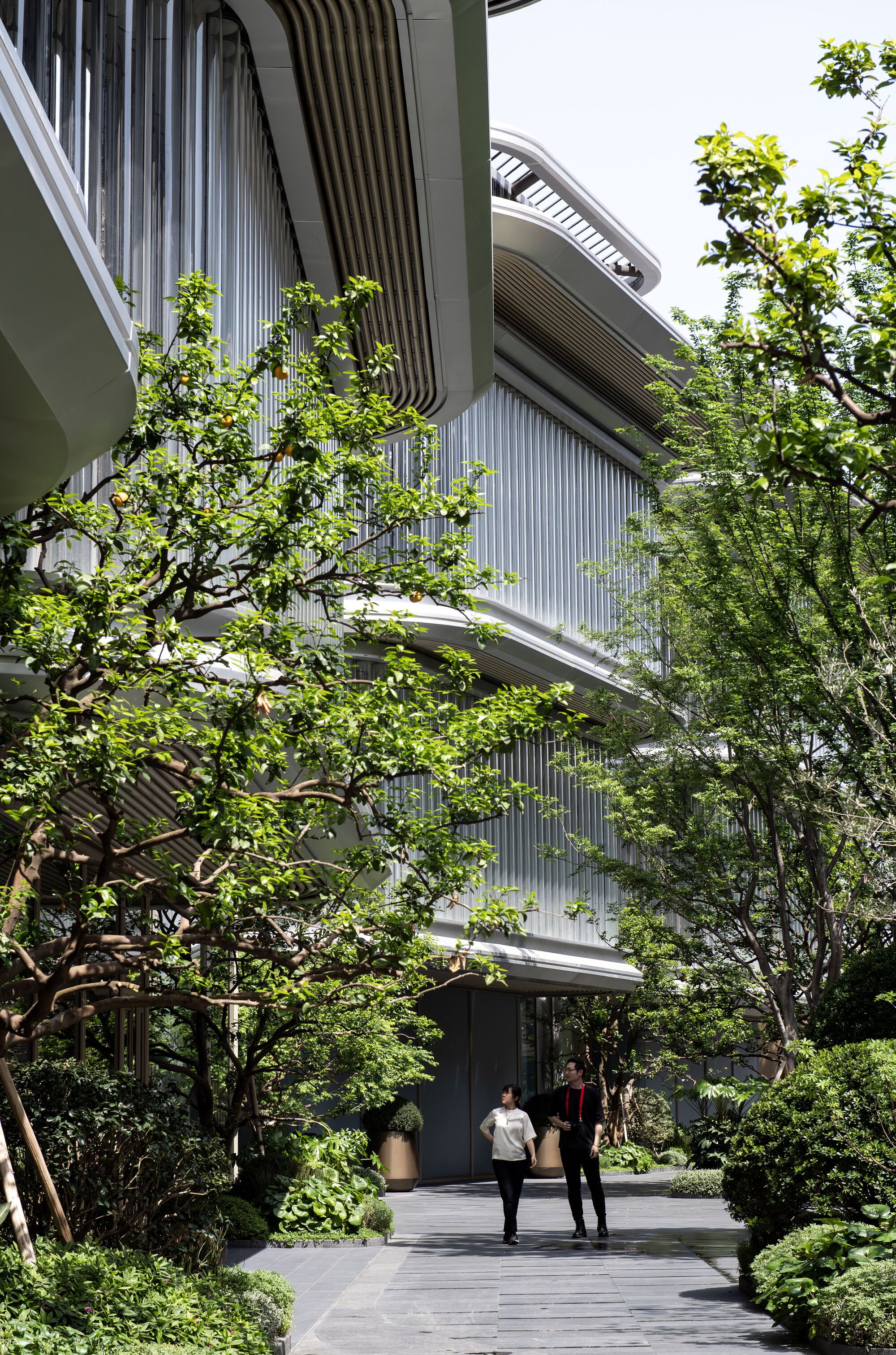Two people walking toward the entrance below stacked balconies wrapped in vertical screens and overhanging branches