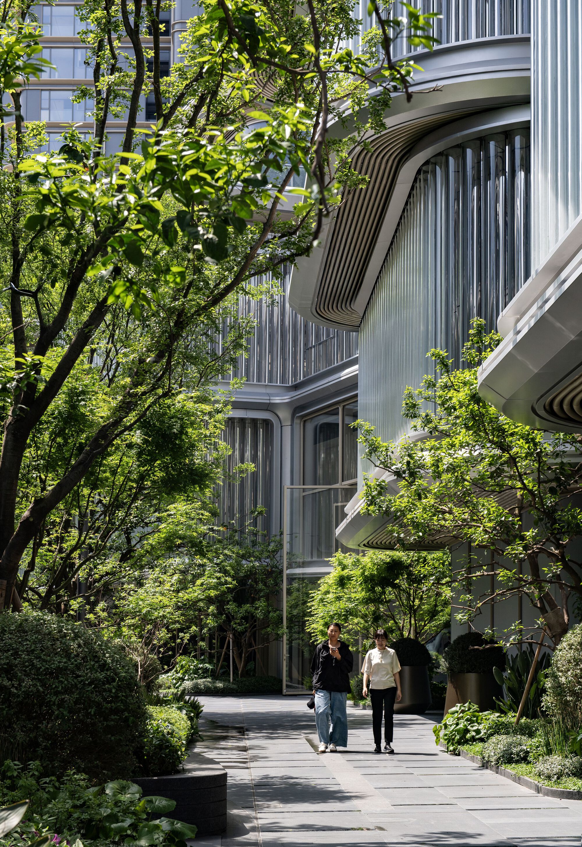 Curved balconies with vertical metal fins framed by mature trees and a landscaped pathway with pedestrians