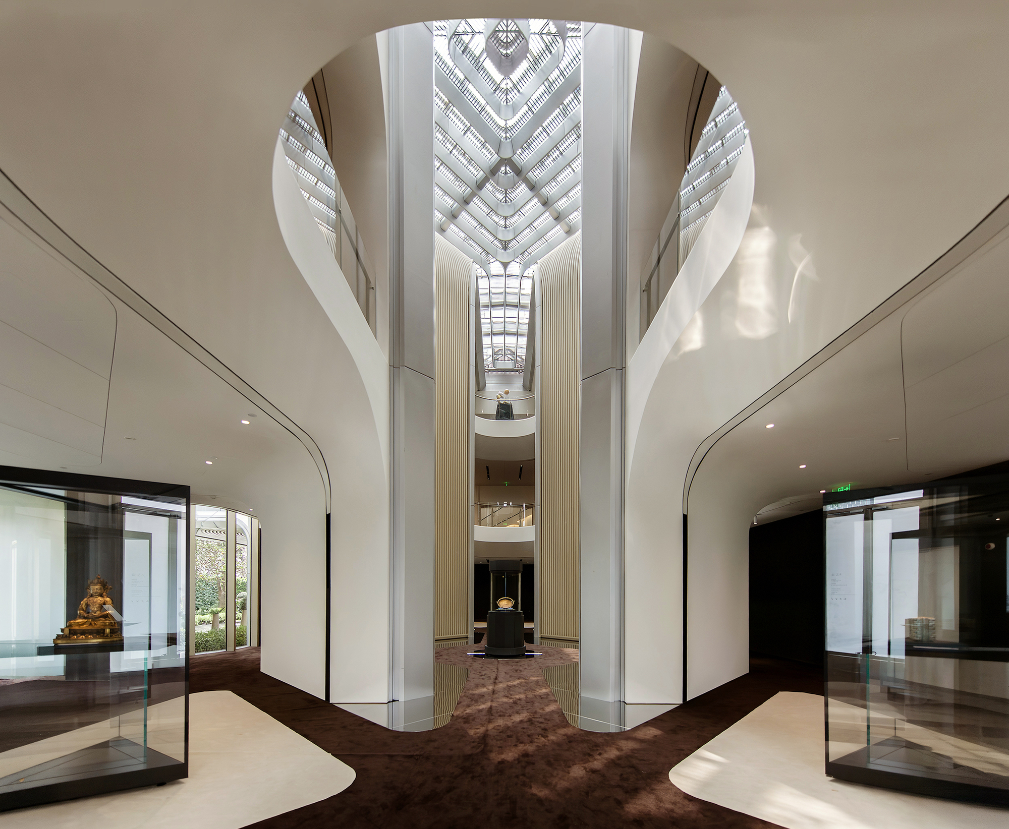 Multi-story atrium with curved white walls and a geometric glass skylight centered above a display platform