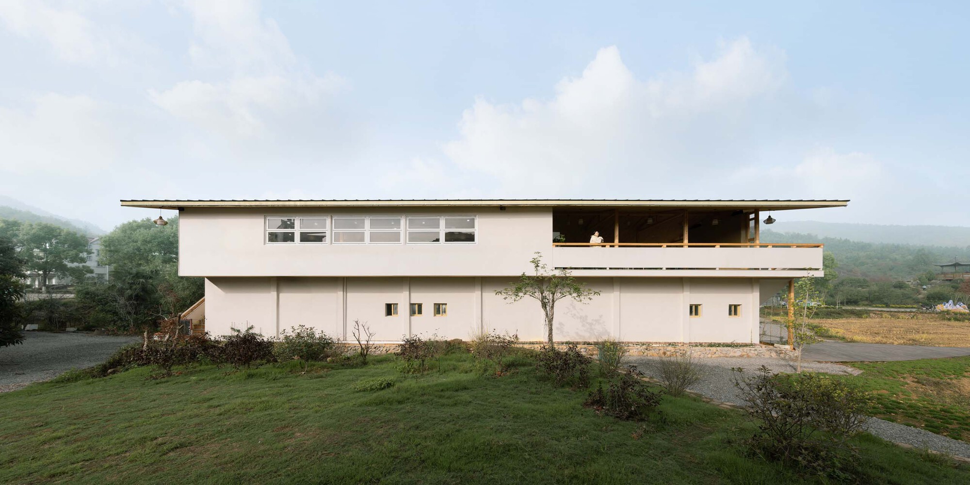 Two-story white stucco residence with overhanging flat roof and upper-level timber deck in a rural valley