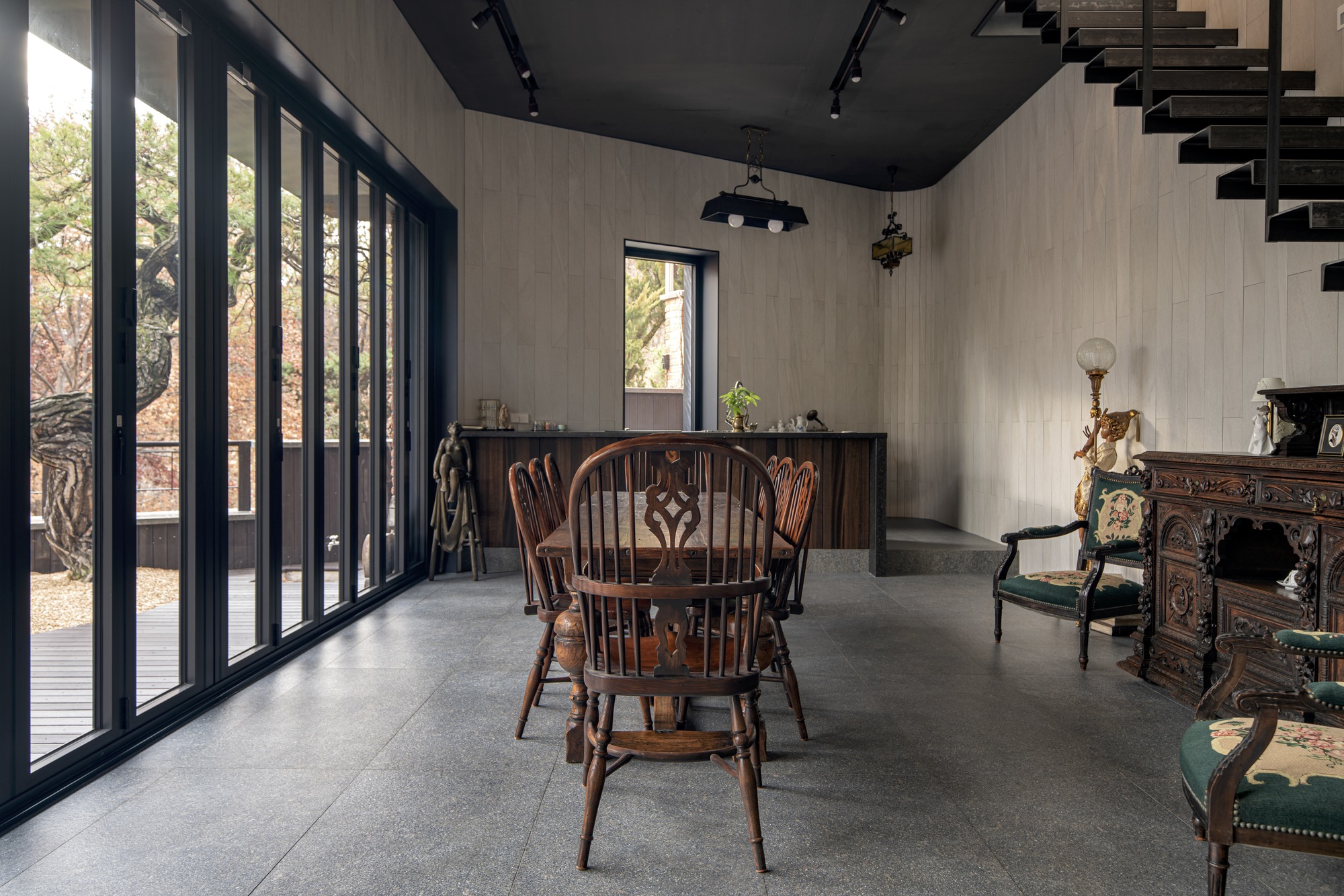 Dining space with tall black-framed glazing overlooking bare trees and a dark ceiling above