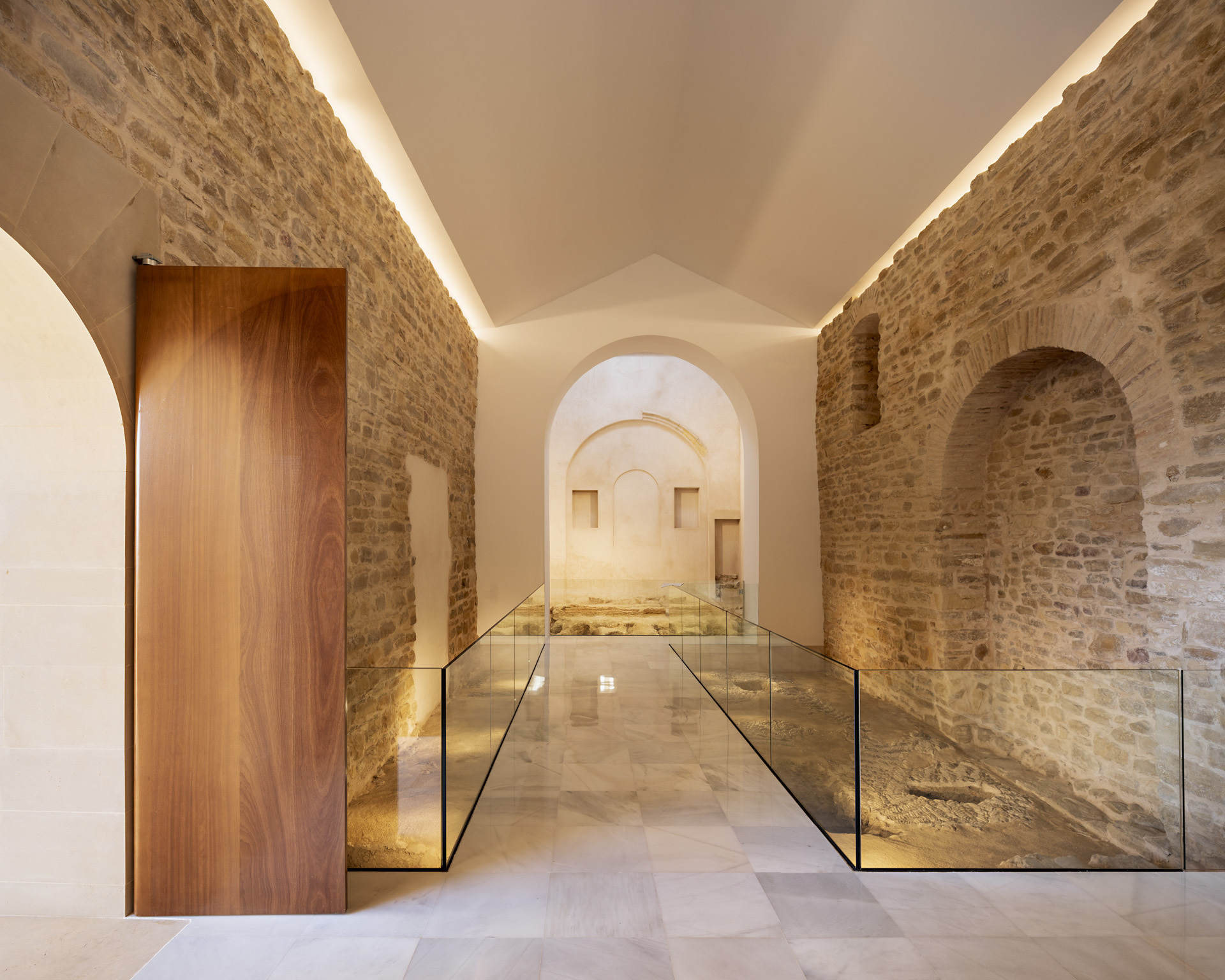 Vaulted corridor with exposed stone walls, walnut door and glass-edged floor revealing archaeological remnants below