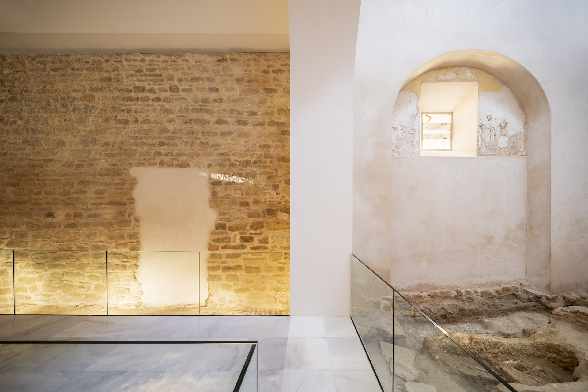 Arched niche in a white plaster wall with glass floor protecting excavated stone foundations beneath