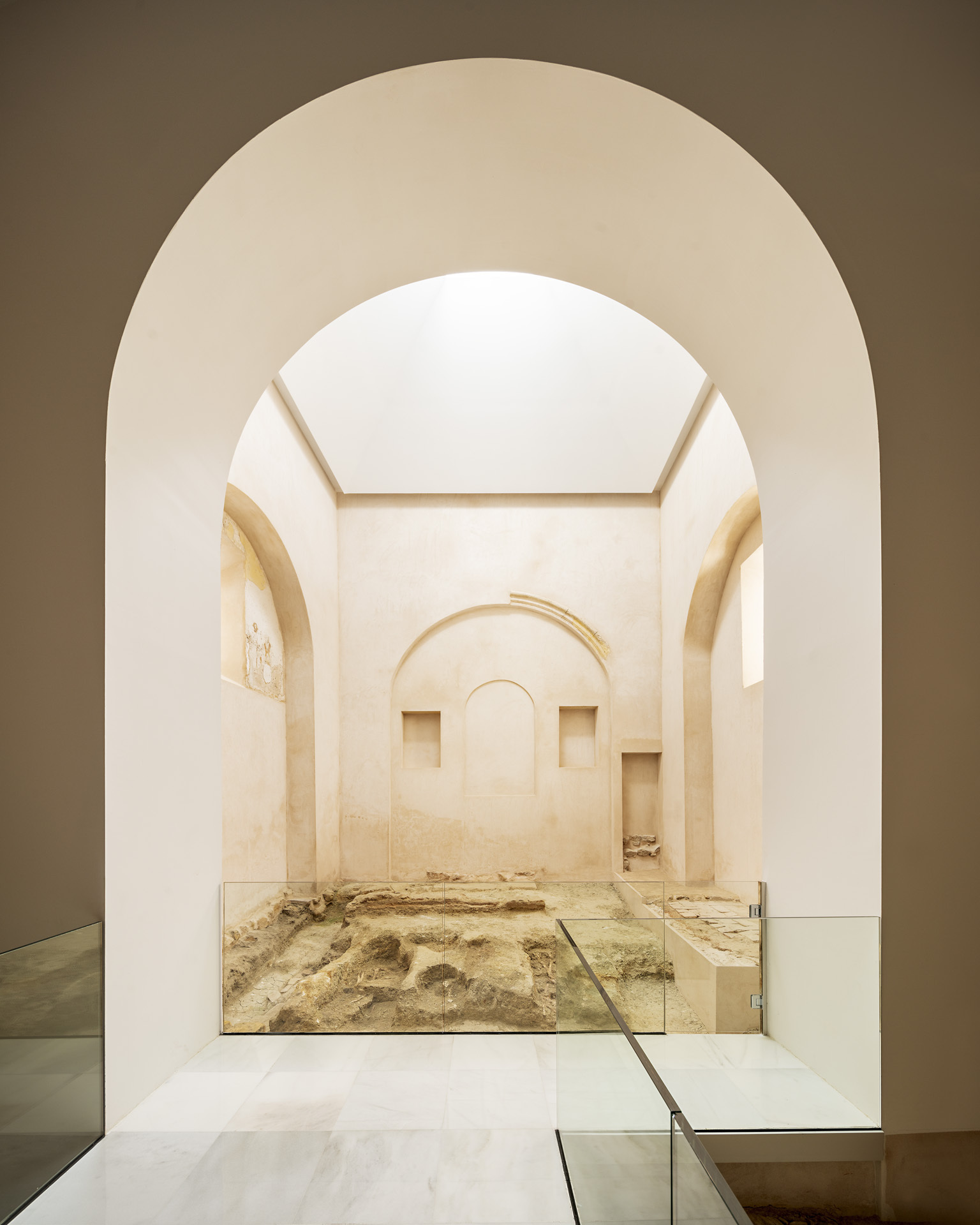 View through layered arches into a whitewashed room with glass-protected archaeological floor and flanking niches