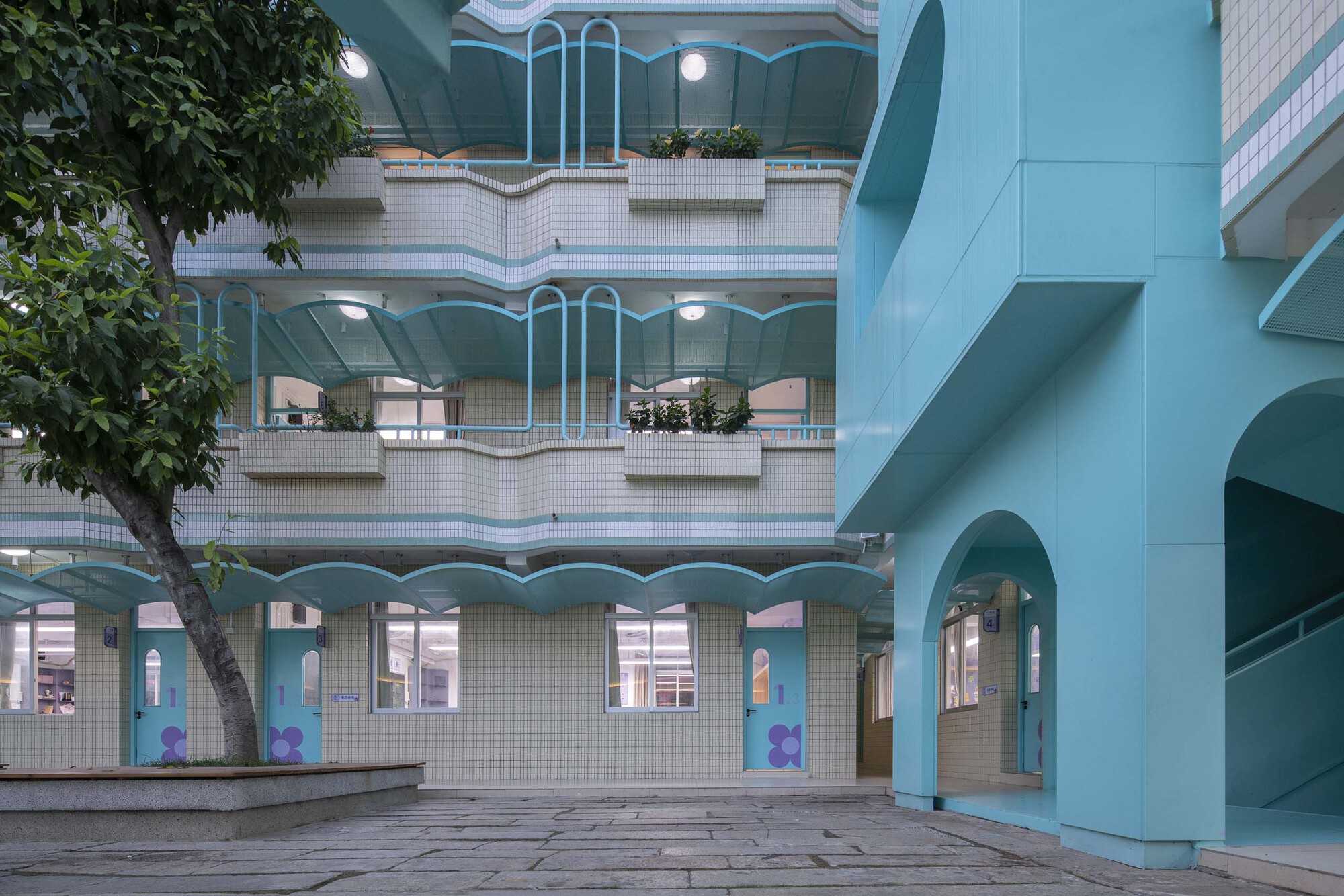 View across a paved plaza showing tiered balconies with wavy white railings and potted trees