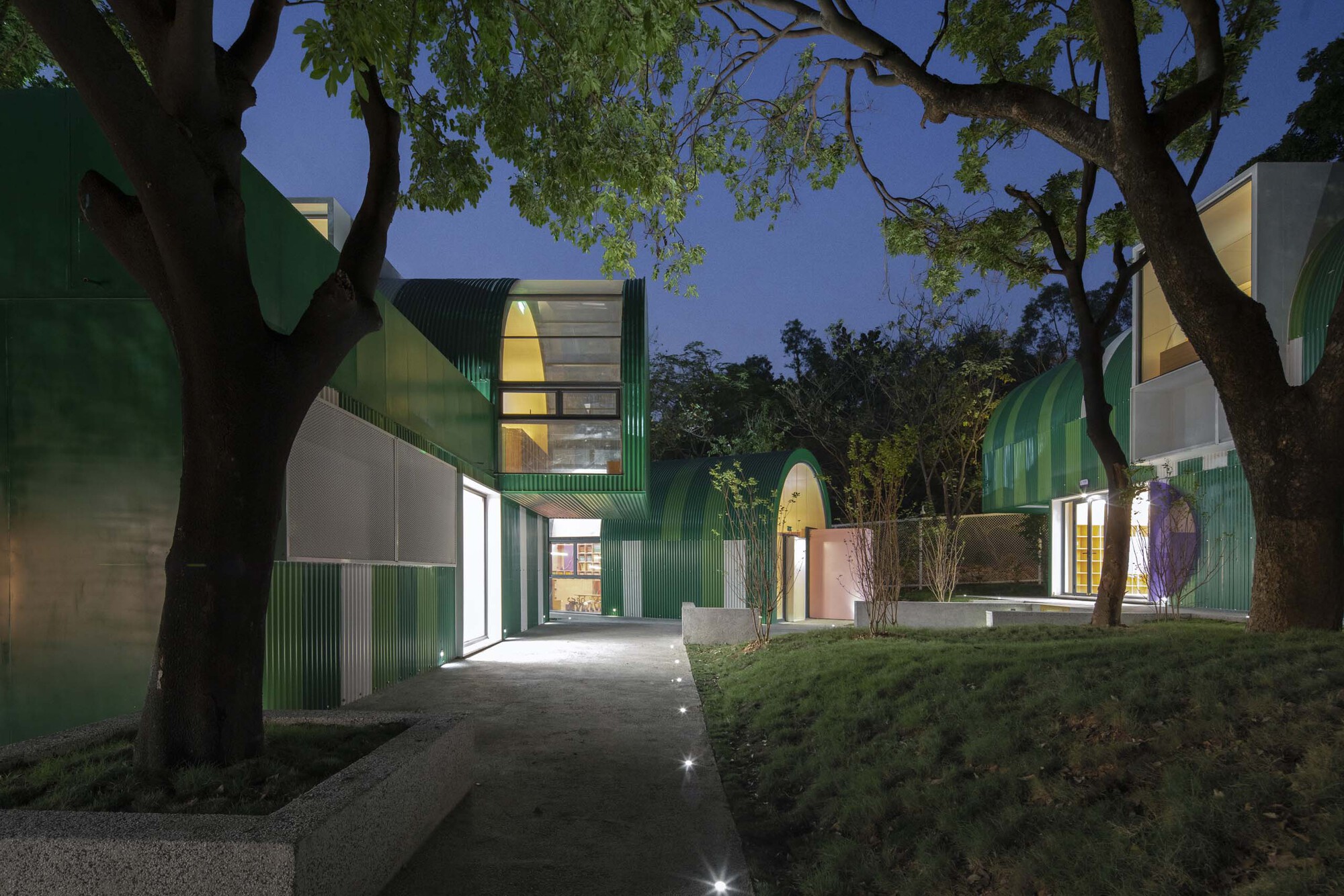 Illuminated pathway between green paneled volumes under mature trees at twilight