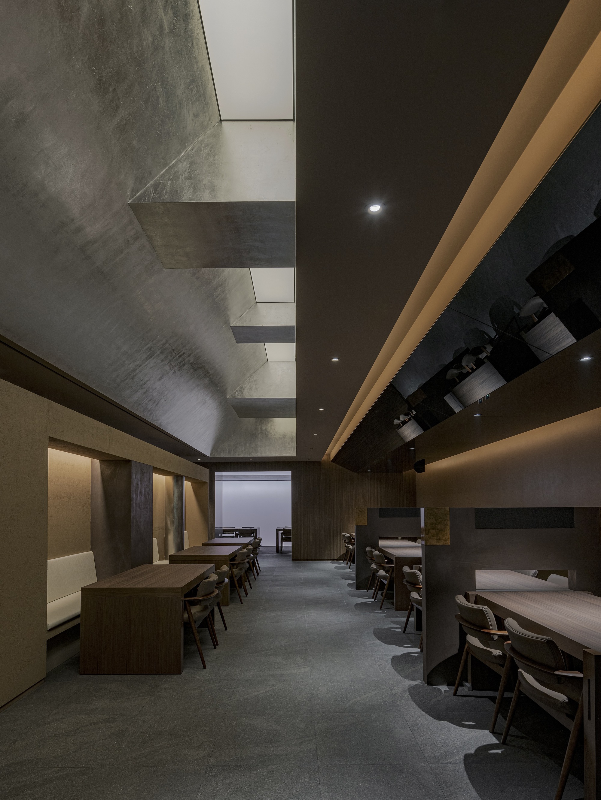 Long dining hall with timber tables, recessed lighting, and a faceted concrete ceiling void above