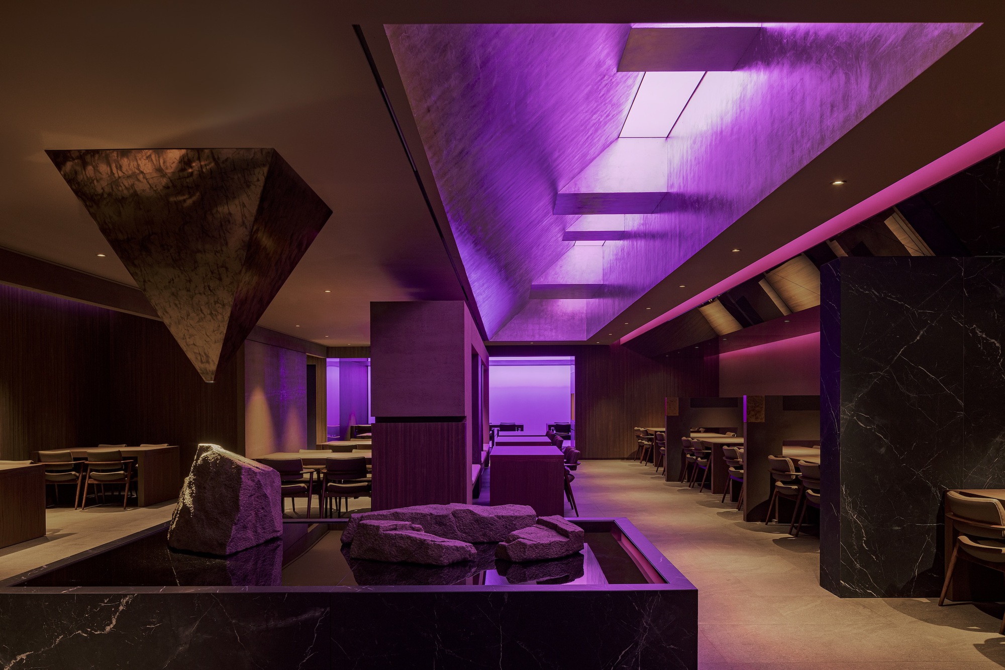 Restaurant dining hall with purple-lit ceiling panels and a rough stone sculpture centerpiece on a black plinth