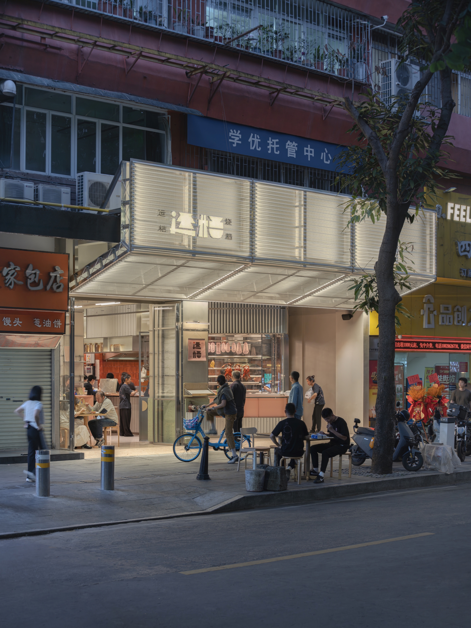 Evening streetscape showing customers gathered outside the shopfront beneath the cantilevered metal canopy