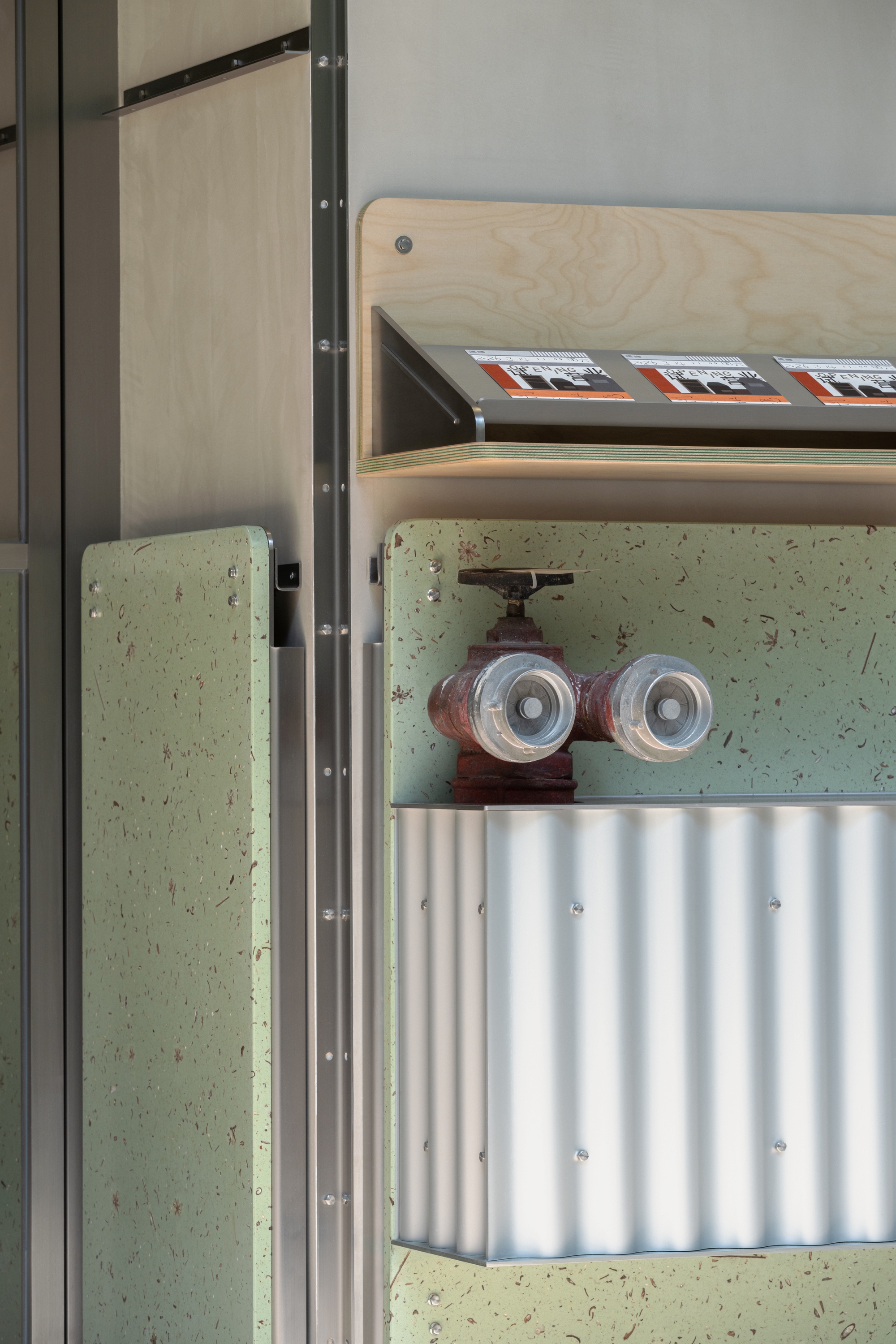 Close-up of exposed water valve and pipes mounted on pale green terrazzo wall beneath plywood shelf
