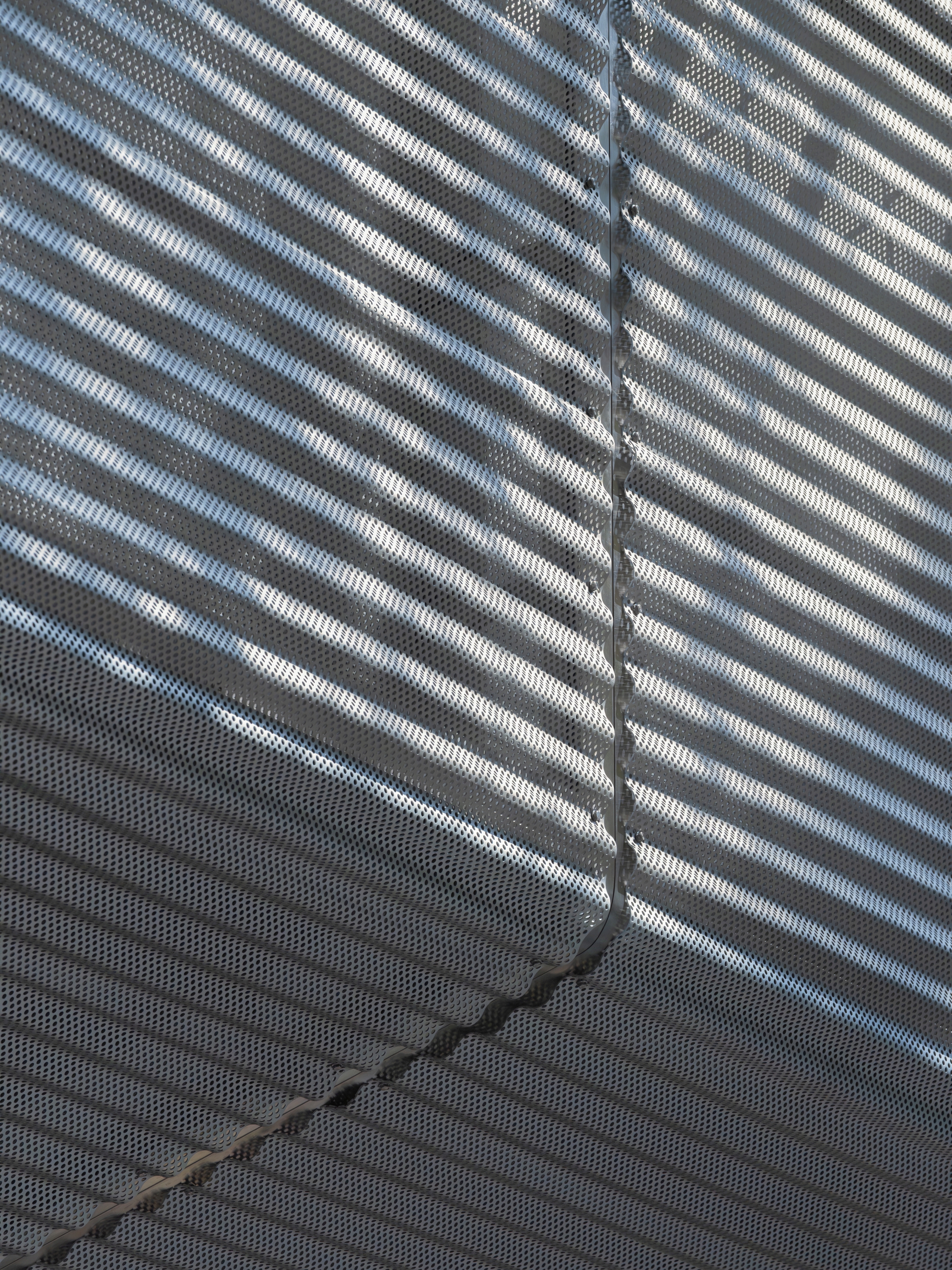 Close-up of perforated metal facade panels with diagonal shadow patterns from afternoon sunlight