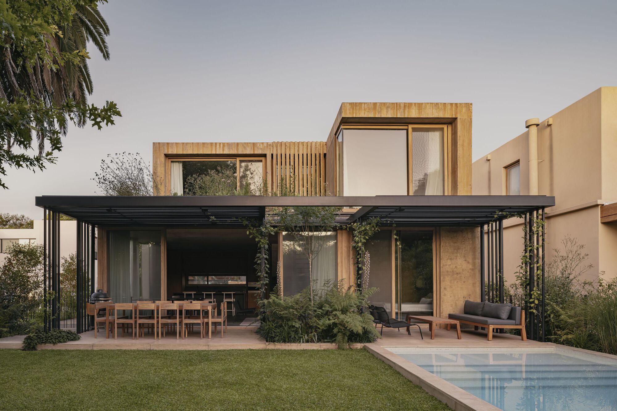 Garden view showing black steel pergola pavilion with outdoor dining and timber-clad upper stories