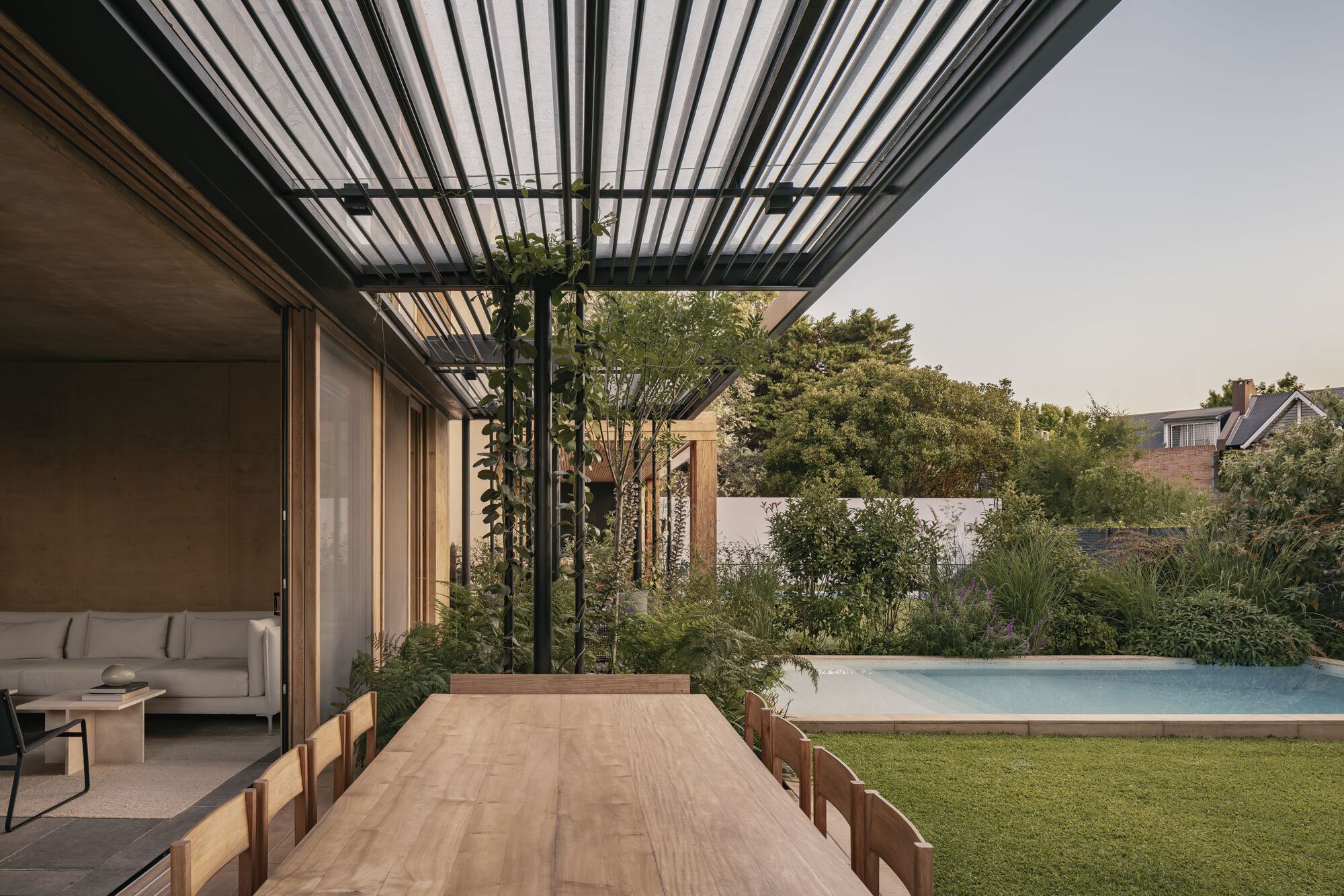 Dining terrace beneath slatted steel canopy with pool and planted beds at twilight