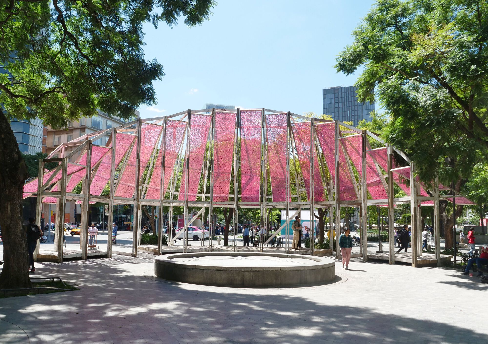 Open timber pavilion with pink fabric roof panels framing a circular concrete fountain under dappled tree shade
