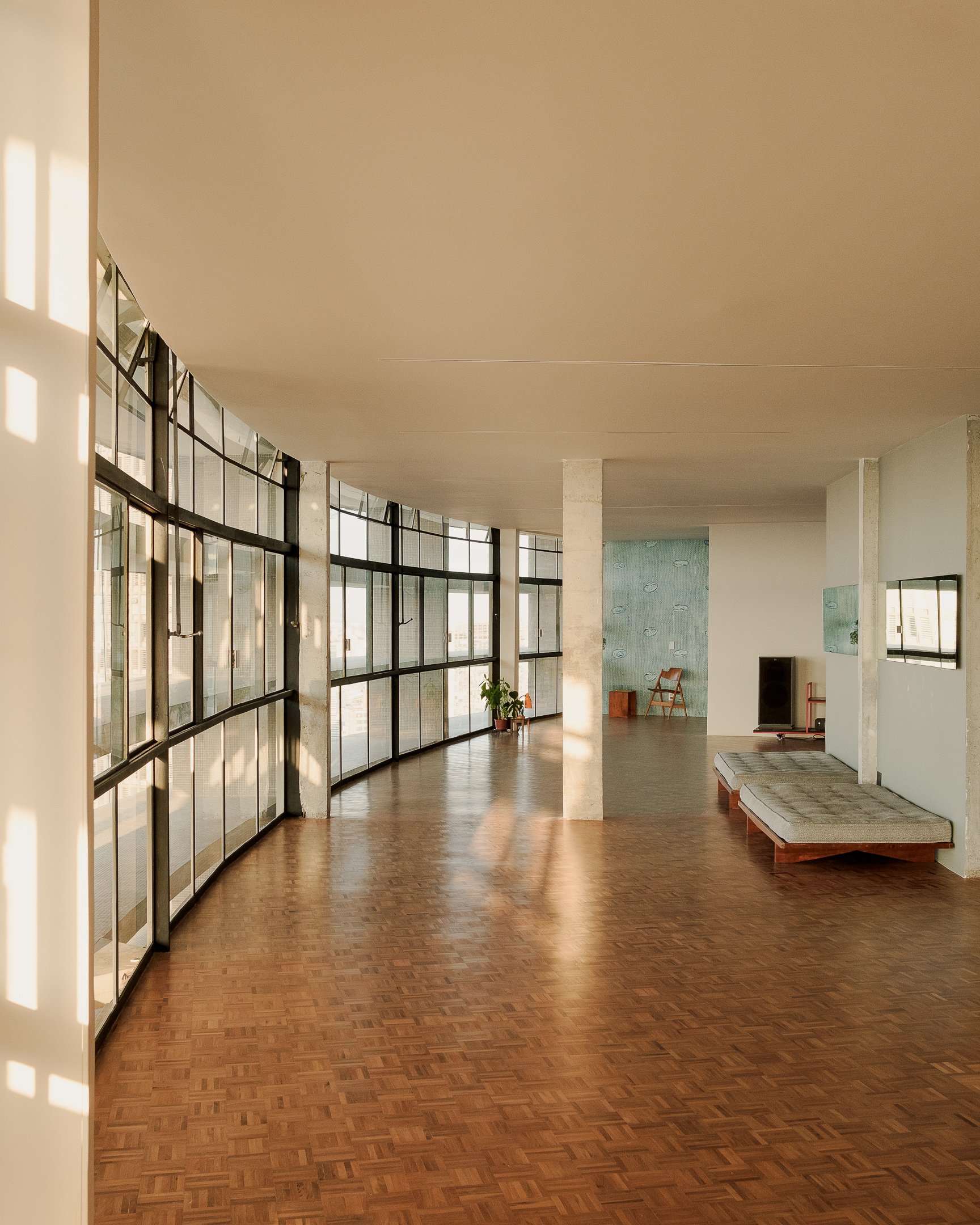 Curved steel-framed glass wall filtering afternoon light into a living area with parquet floor and low bench