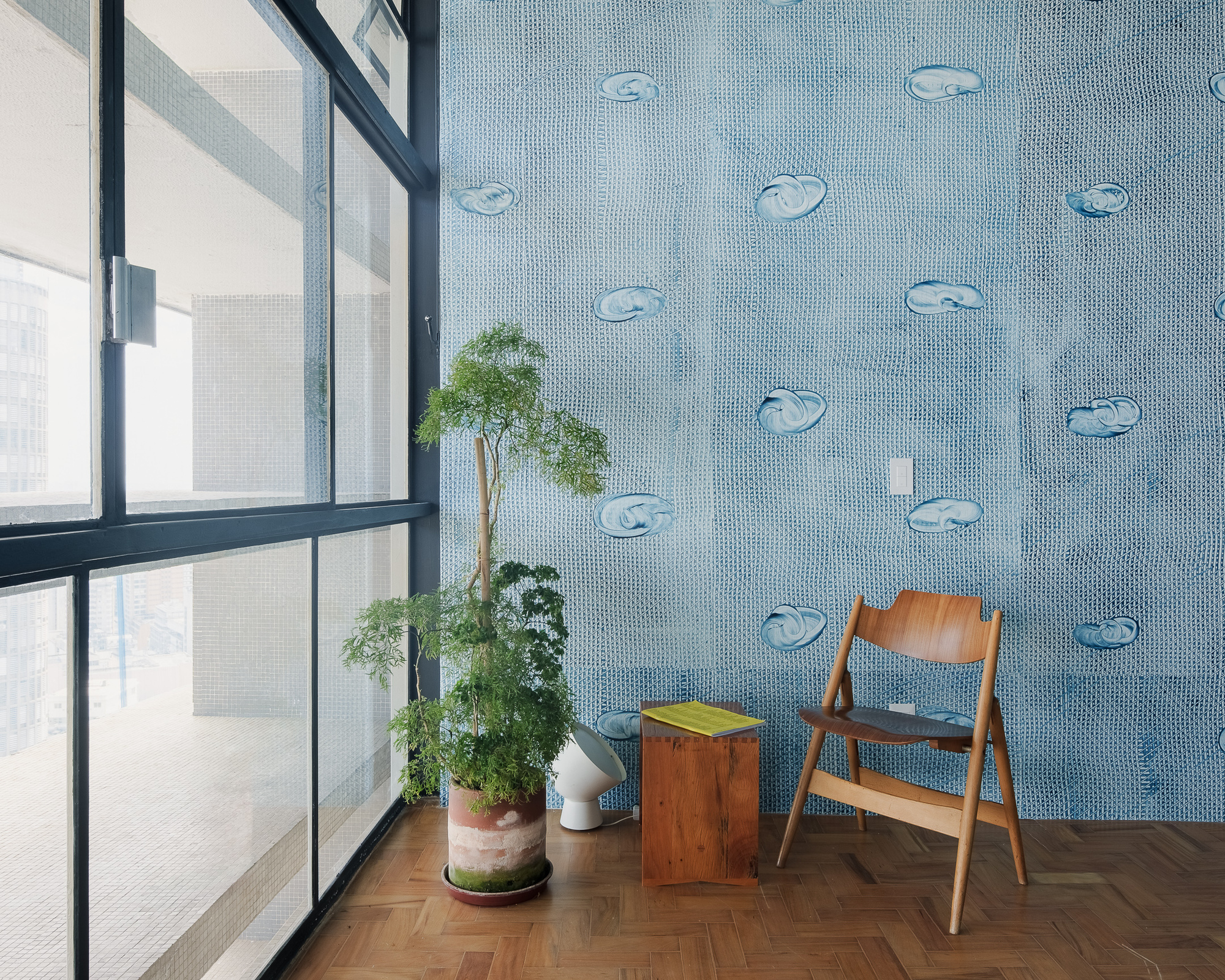 Blue mosaic tile wall with fish motifs beside a steel-framed window and wooden chair with potted plants
