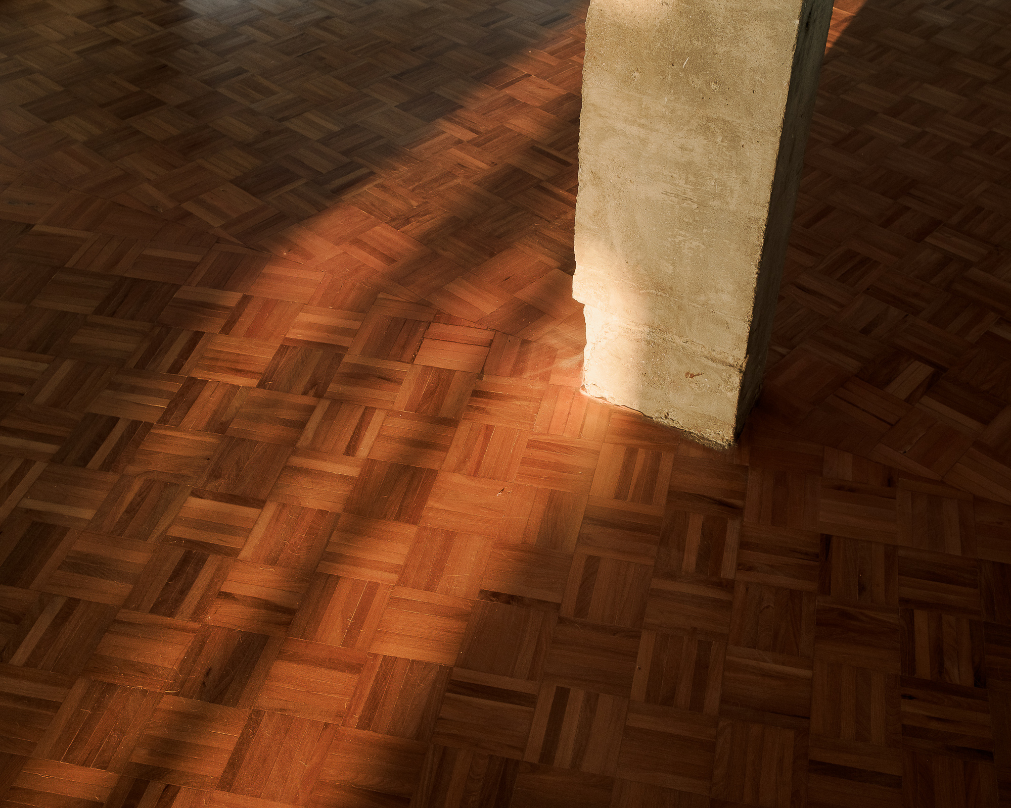 Herringbone parquet floor meeting a textured concrete column base in warm afternoon sunlight