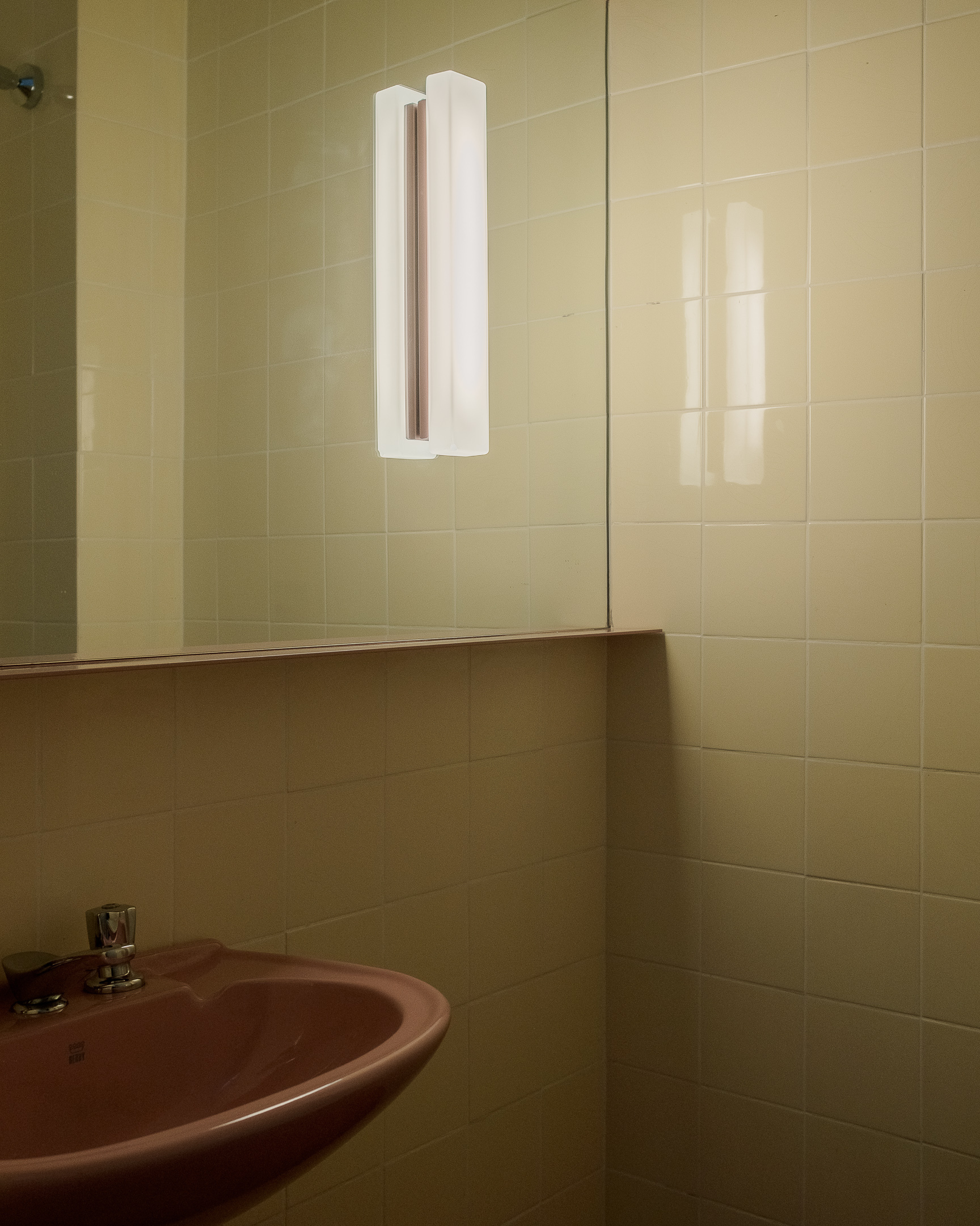 Yellow tiled bathroom corner with mirror and wall-mounted tubular light fixture above a pink sink