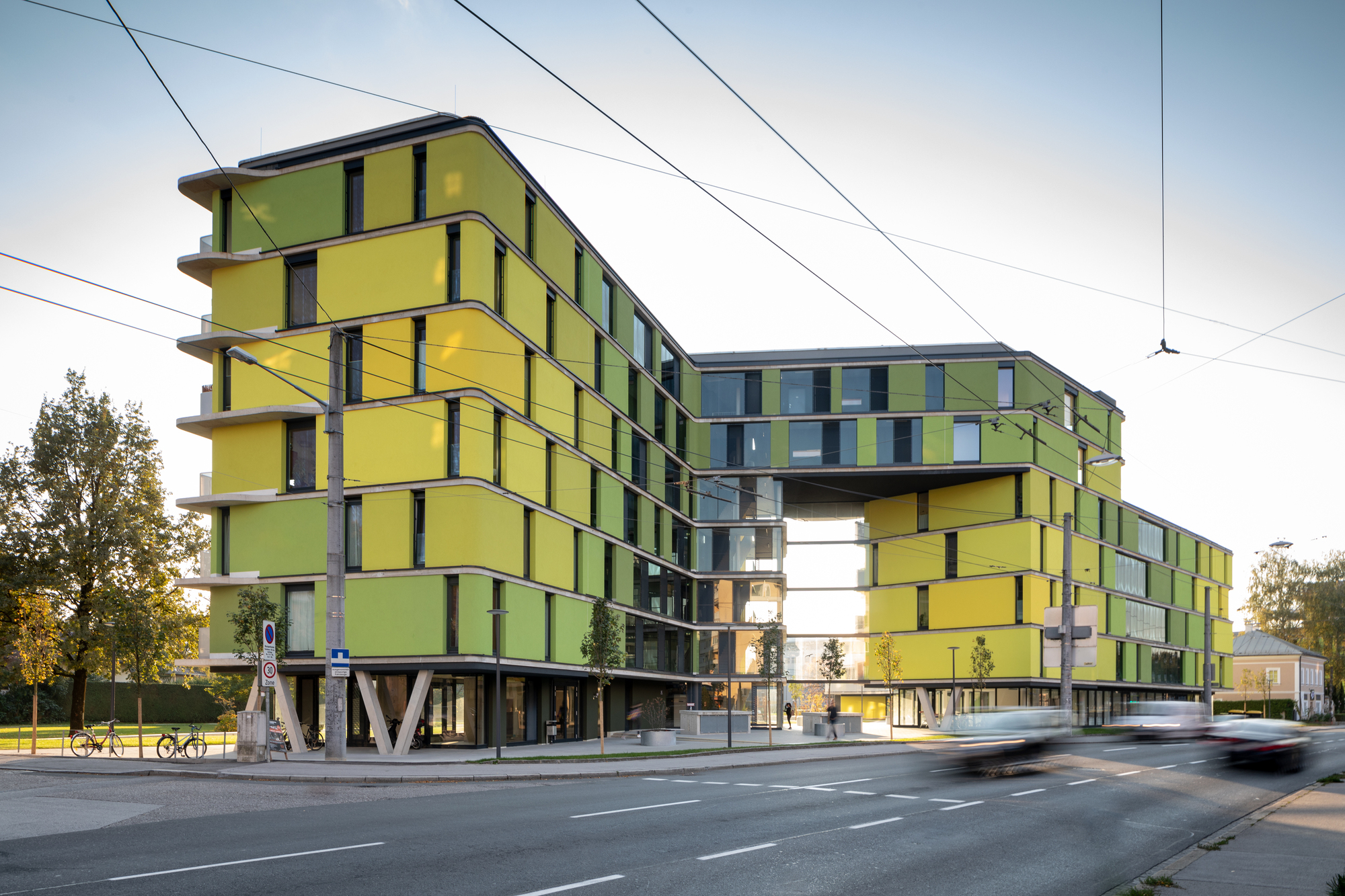 Streetside view of the yellow and green panelized facade on pilotis with overhead tram wires at dusk