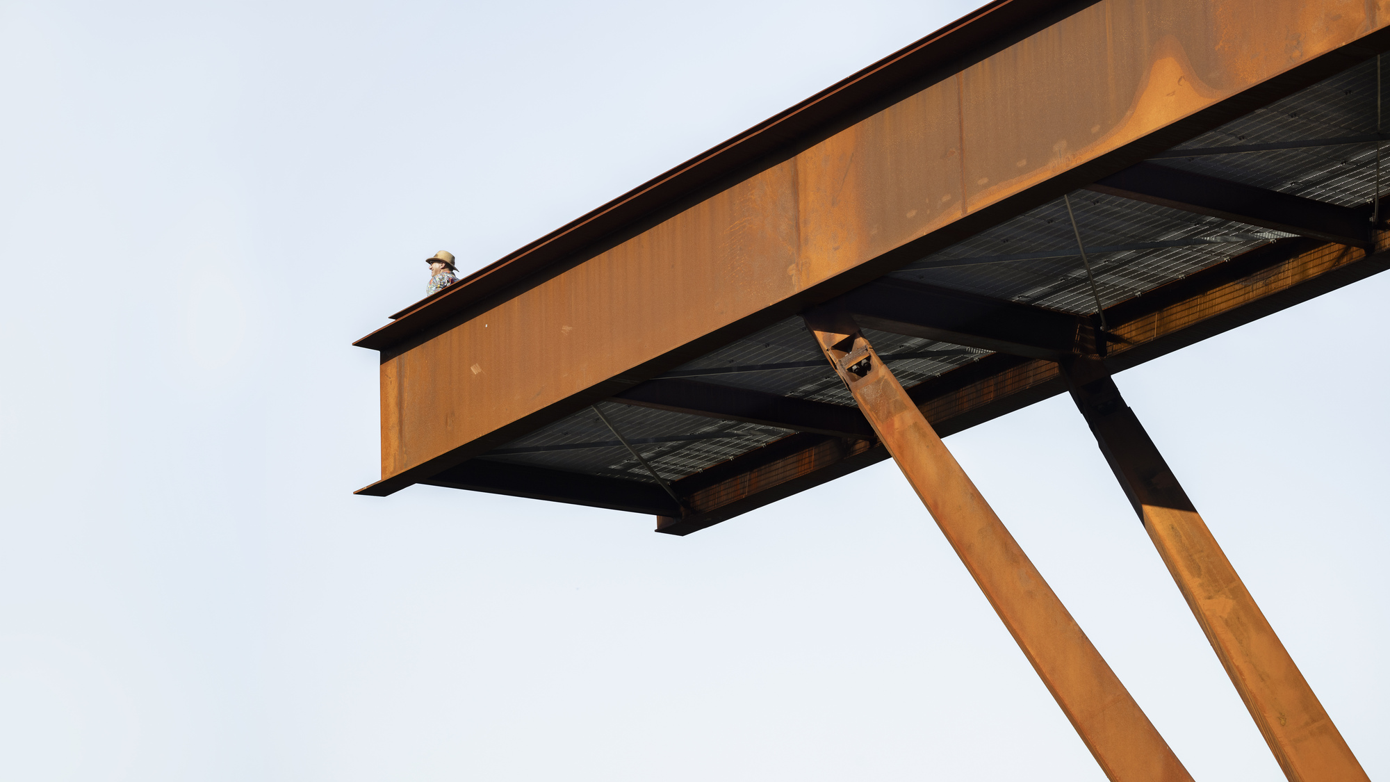 Underside view of the cantilevered steel beam supported by angled weathering steel columns against an overcast sky