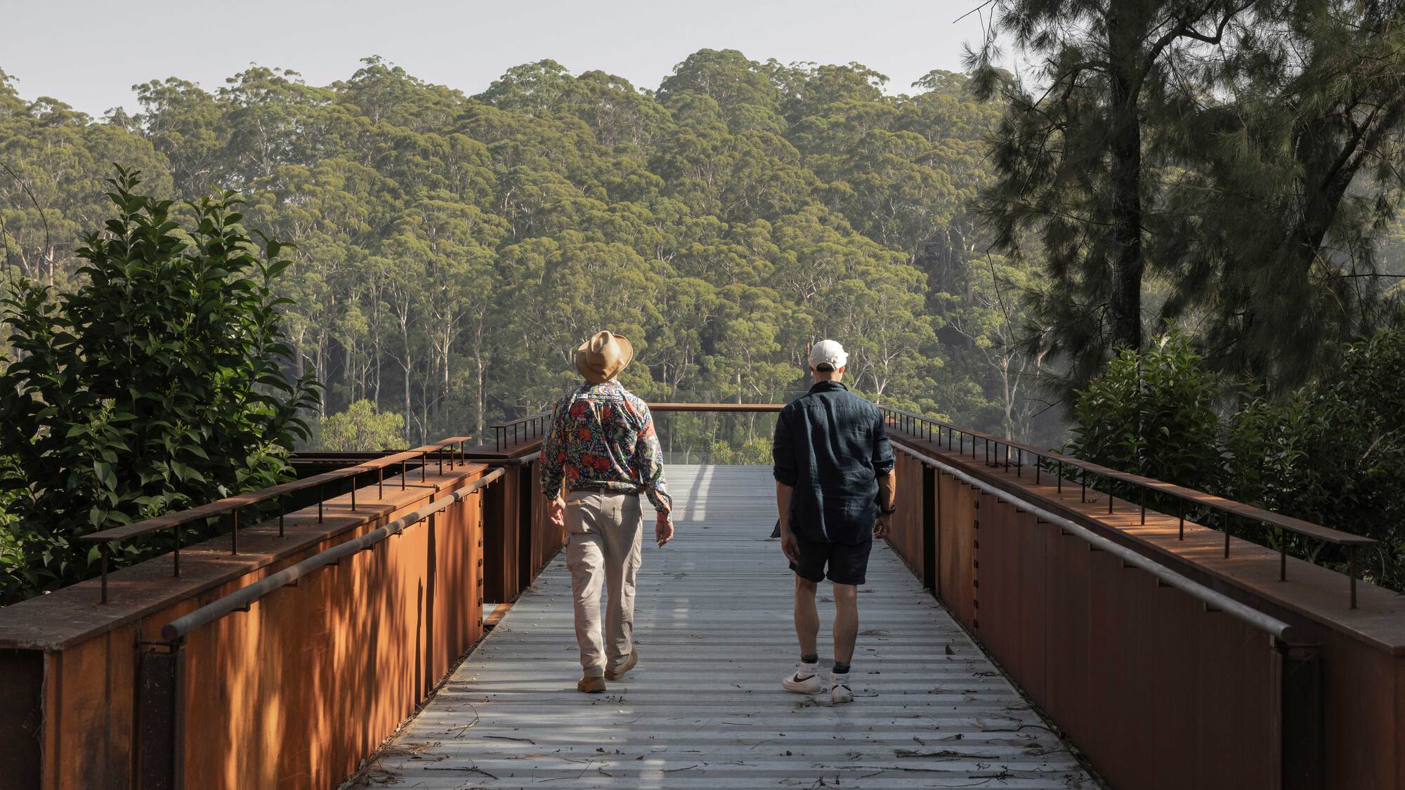 Two visitors walking across the weathering steel pedestrian bridge with forest canopy in the background