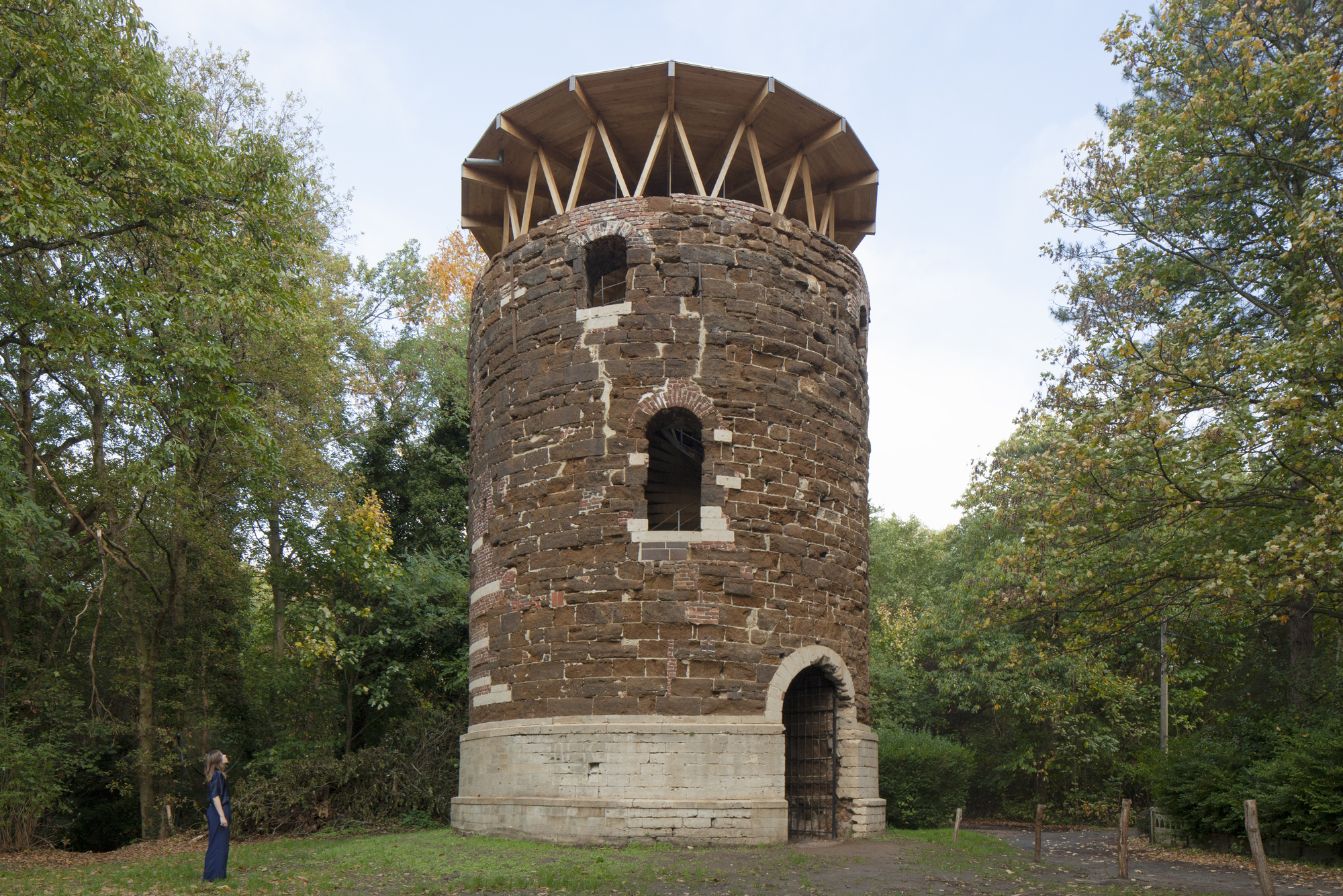 Cylindrical brick tower with zigzag timber roof structure framed by trees on a grassy clearing