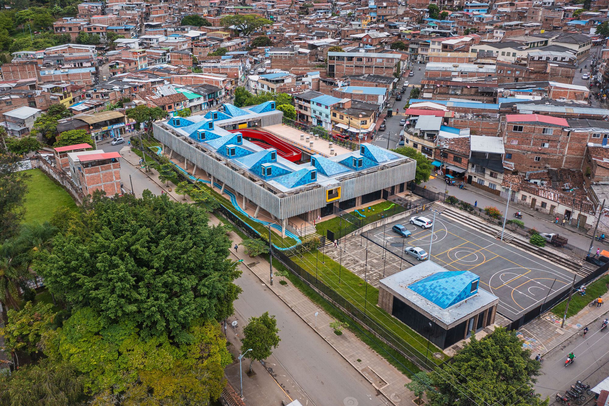Drone view showing the linear building with blue sawtooth roofs and red central courtyard amidst the dense urban fabric