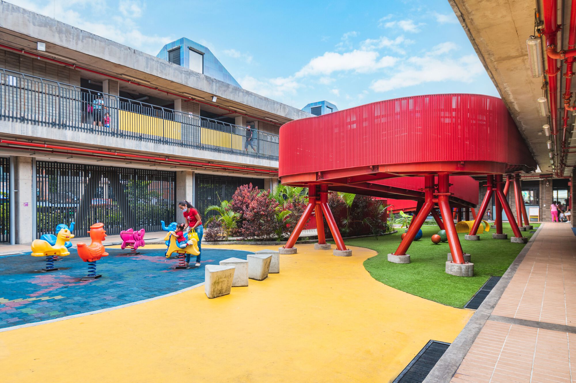 Children playing in a courtyard with blue wading pool and yellow rubber surfacing beneath the curved red pedestrian bridge