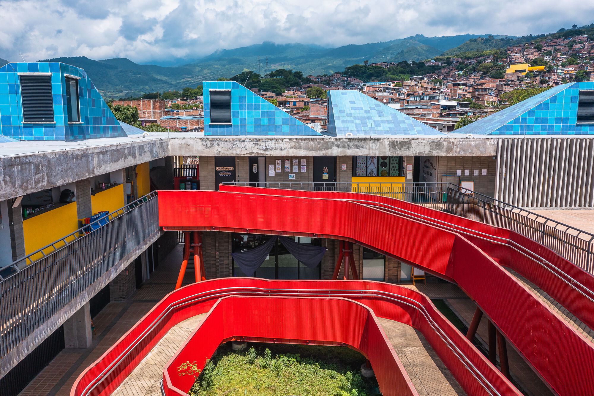 Aerial view of red curved pedestrian bridges connecting concrete buildings with blue pyramid roofs against a mountainous backdrop
